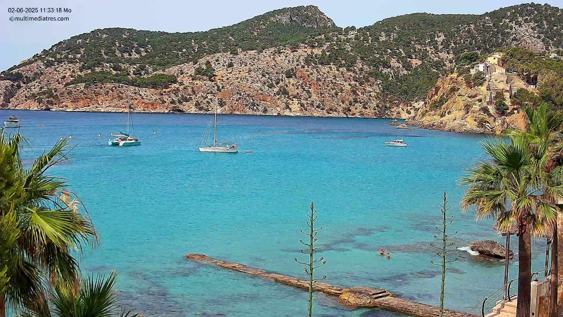 A picturesque cove with turquoise water, several sailboats, and palm trees, under a clear sunny sky, is nestled against a rocky, green hillside.