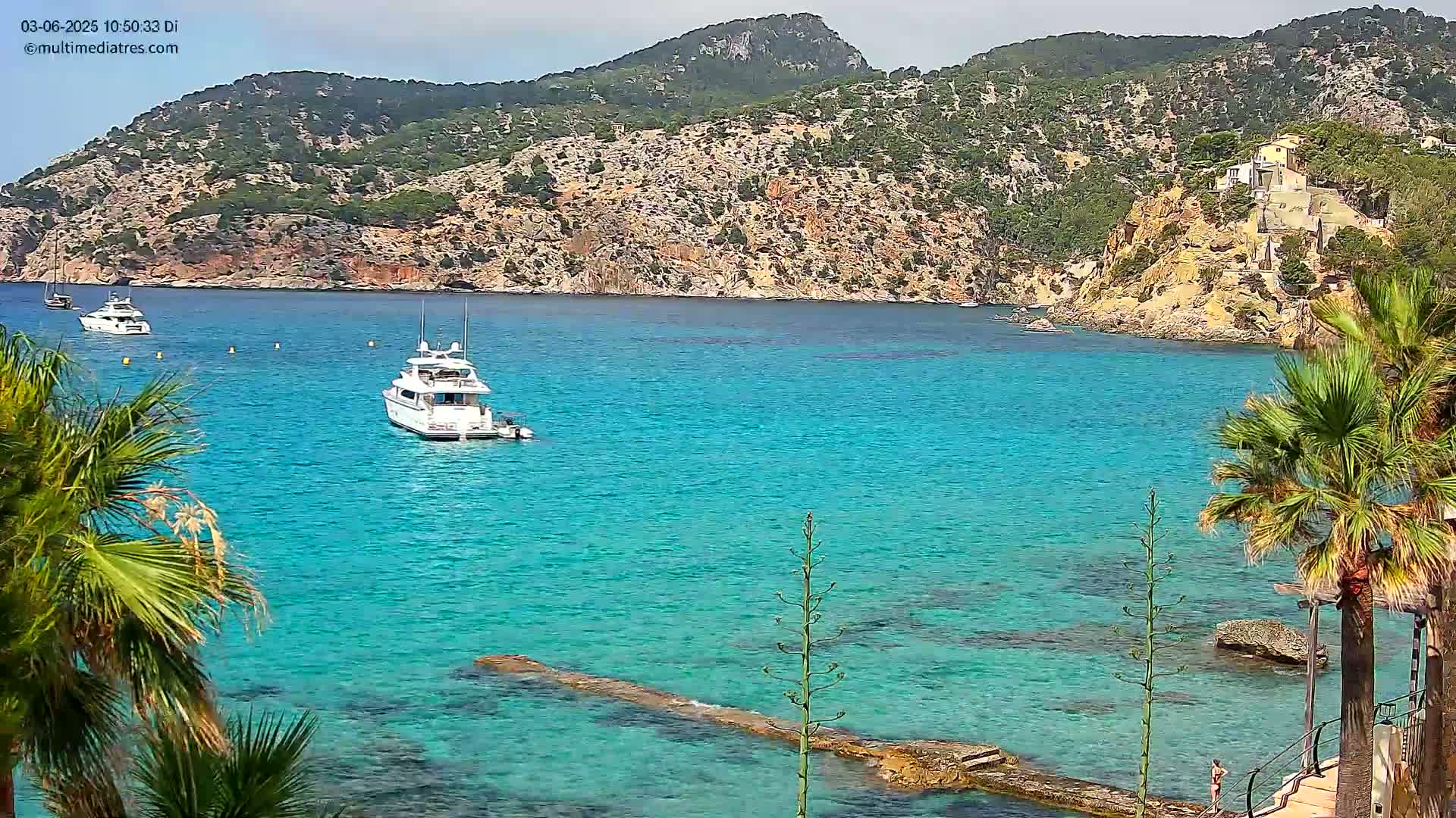 Turquoise water laps a rocky shore, where two yachts are anchored near a verdant, mountainous coastline under a mostly clear sky.