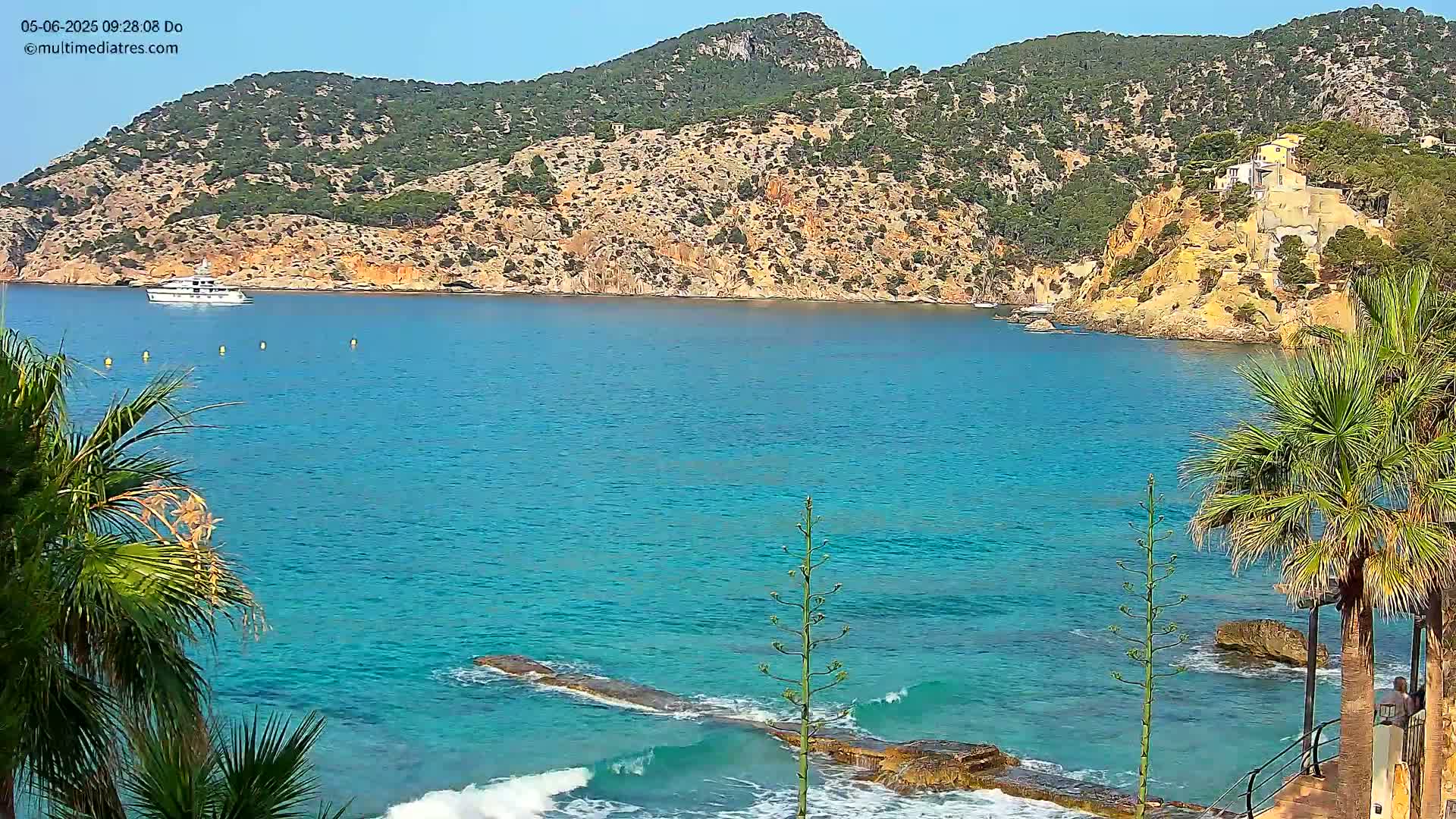 A yacht is visible on a calm, bright, turquoise ocean next to a rocky, green-covered hill with small buildings, framed by palm trees on the shore.
