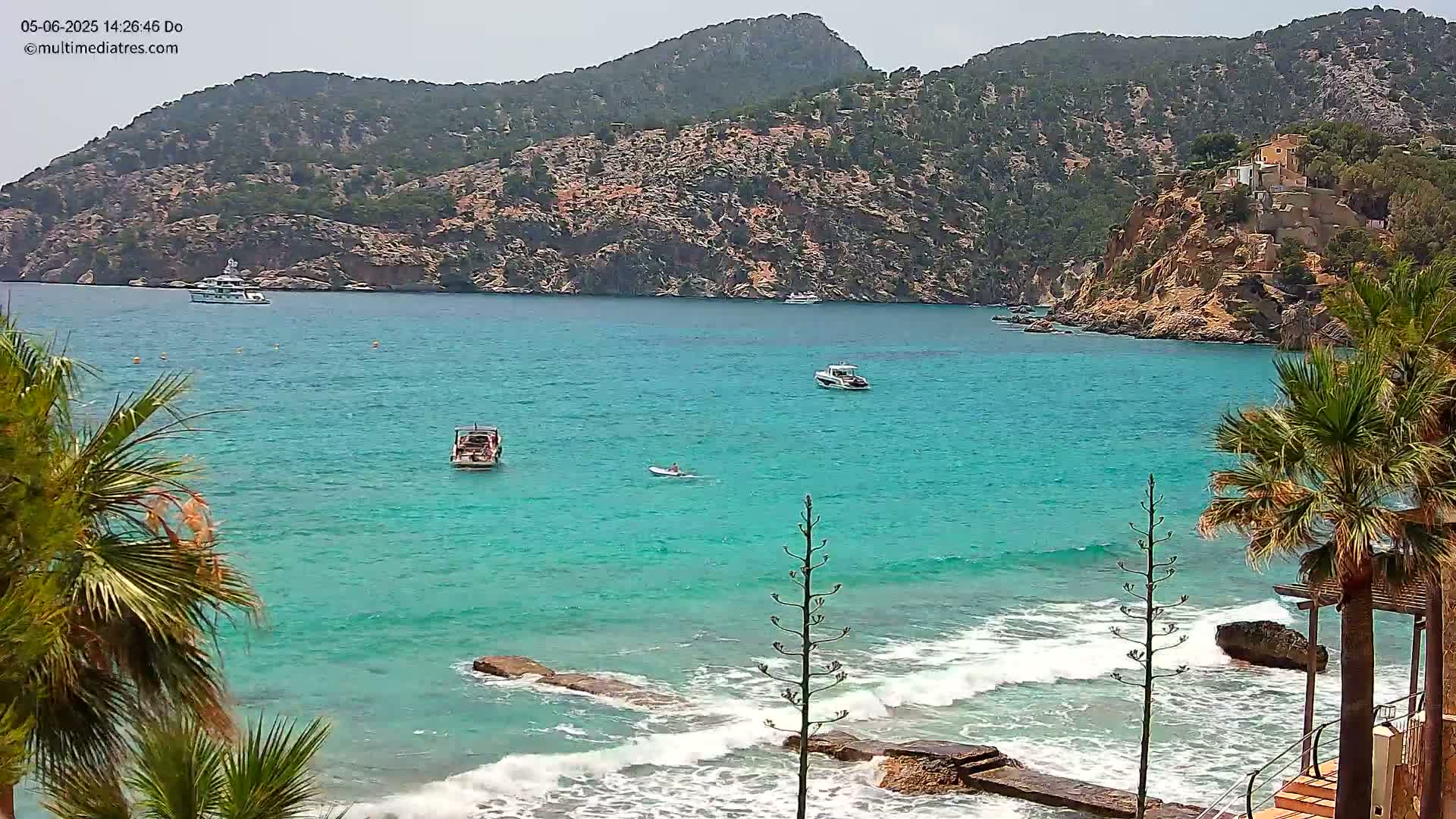 A clear, sunny day at a turquoise bay, featuring several small boats, a person paddleboarding, rocky cliffs, and palm trees along the shore with small waves breaking on the rocks.