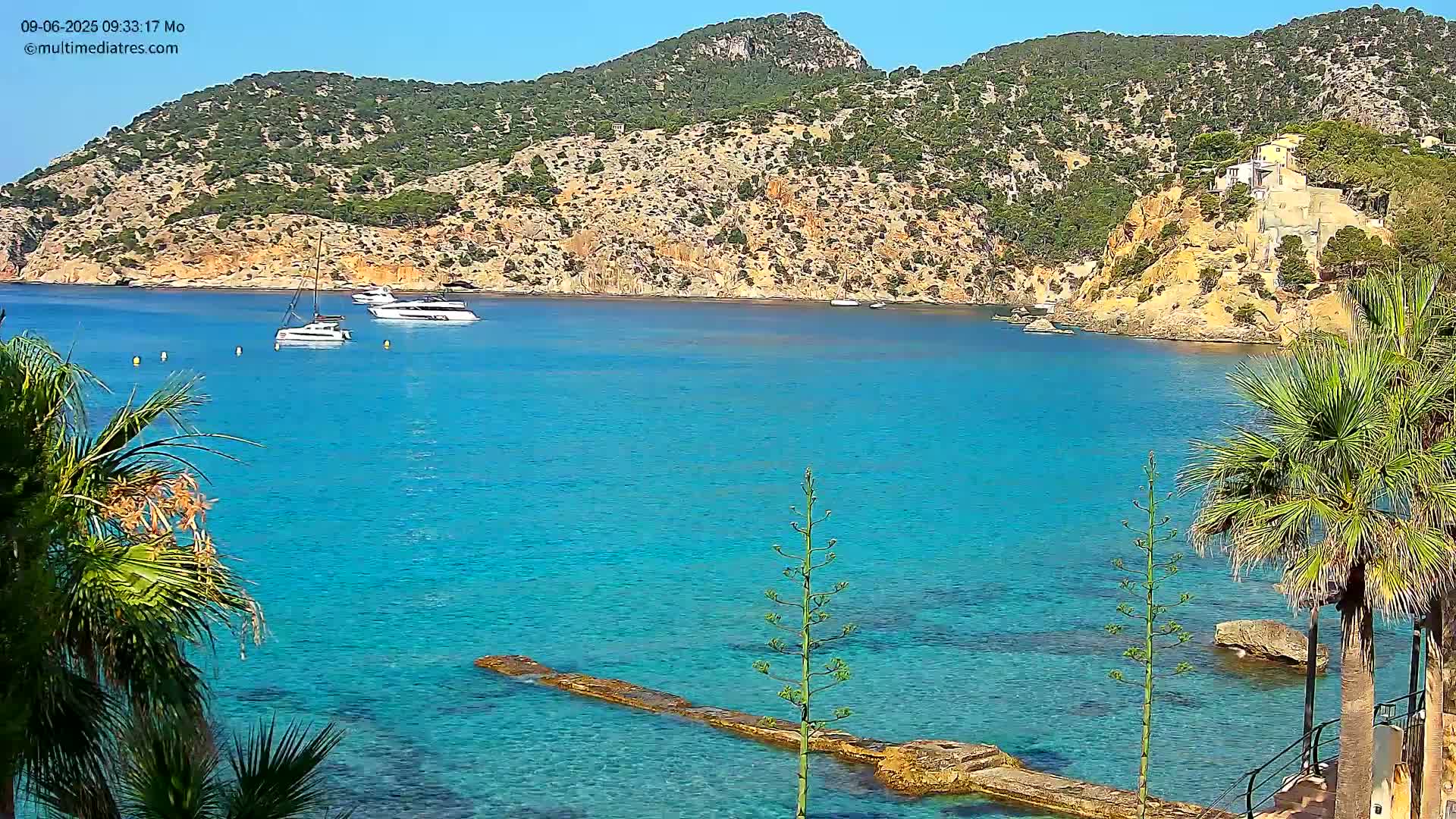 A tranquil, turquoise bay is nestled against a rocky, green hillside under a clear blue sky, with several boats anchored in the water and palm trees framing the view.