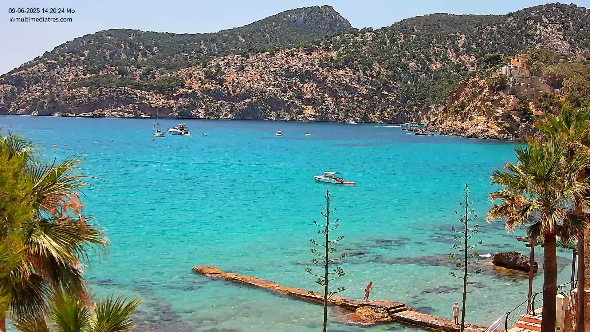 A clear, sunny day reveals a tranquil turquoise bay nestled between rocky hills, with several boats and a few people visible near a low-lying stone pier.