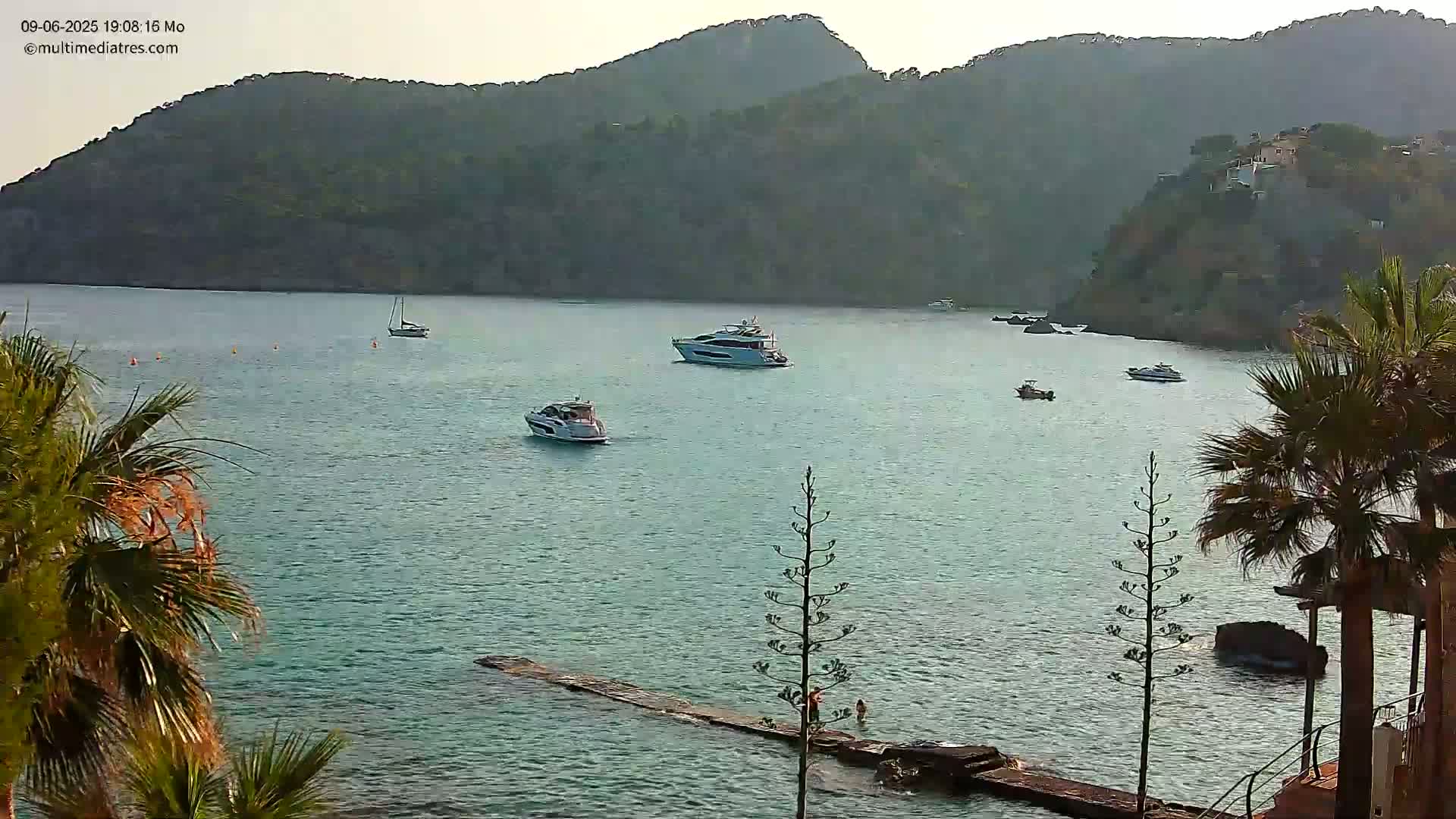 Several boats are moored in a calm, turquoise bay surrounded by dark green hills under a hazy sky.