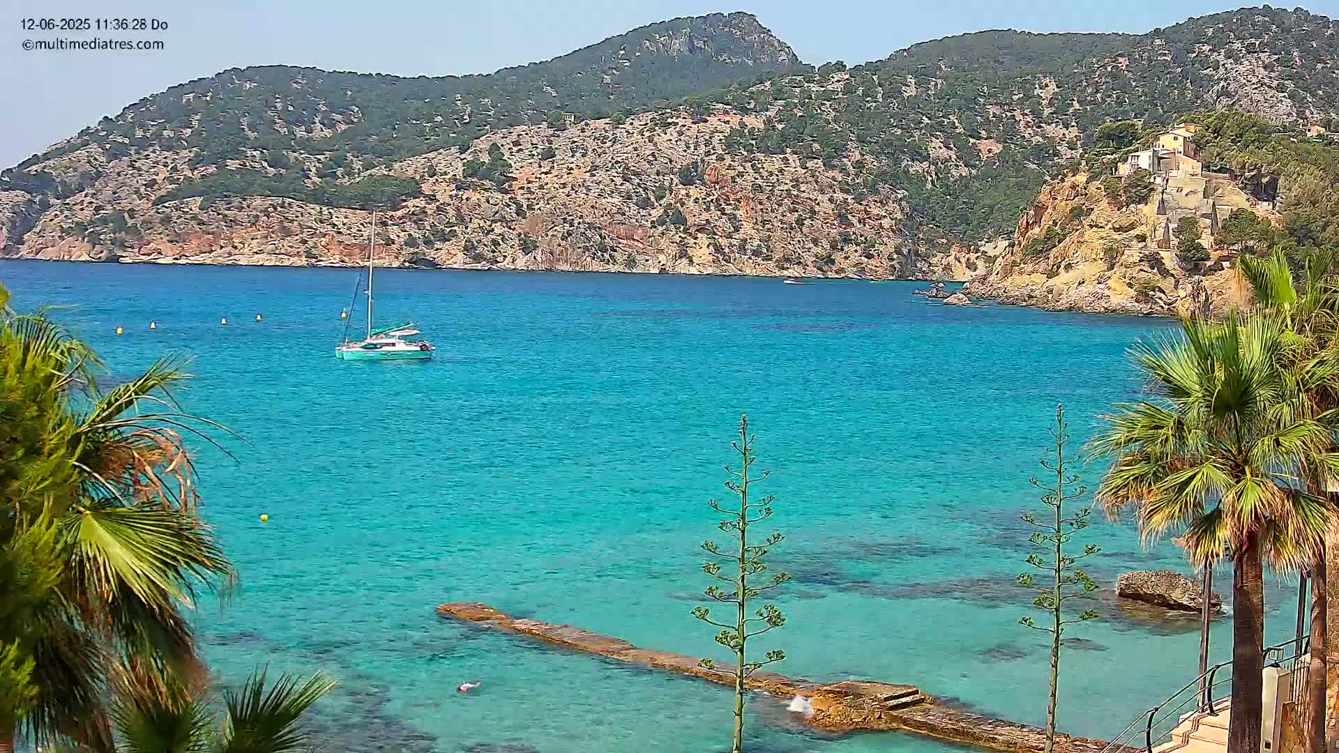 A sailboat is anchored in a calm, turquoise bay surrounded by rocky hills and palm trees on a sunny day.