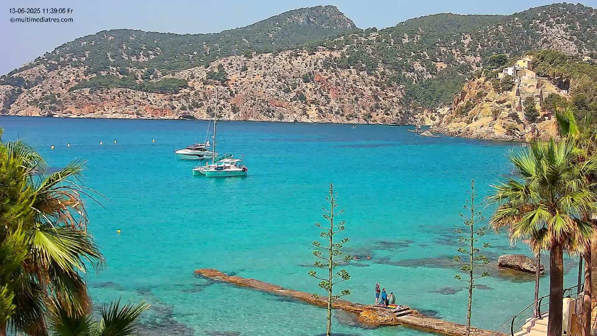 Two sailboats are anchored in a calm, turquoise bay surrounded by a rocky, hilly coastline dotted with palm trees and a few people stand on a low stone jetty.