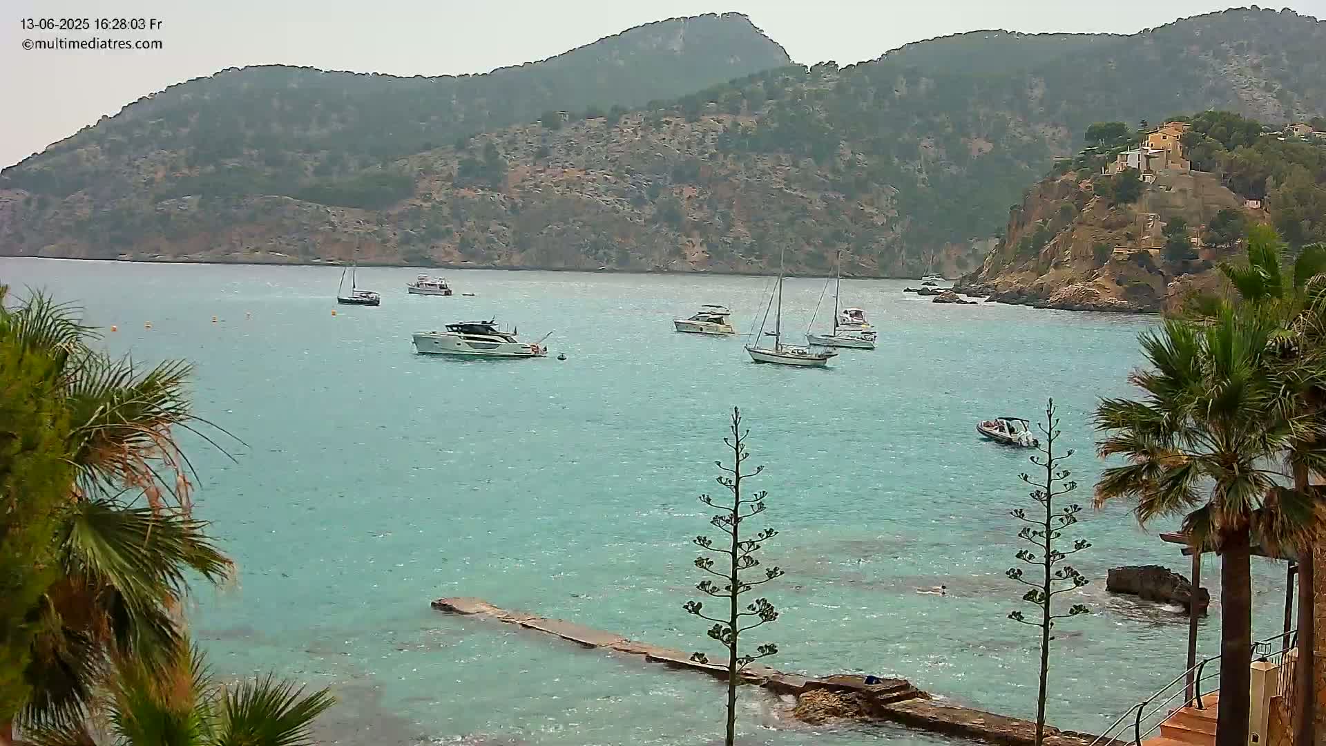 Several boats are anchored in a calm, turquoise bay surrounded by rocky hillsides and palm trees under an overcast sky.