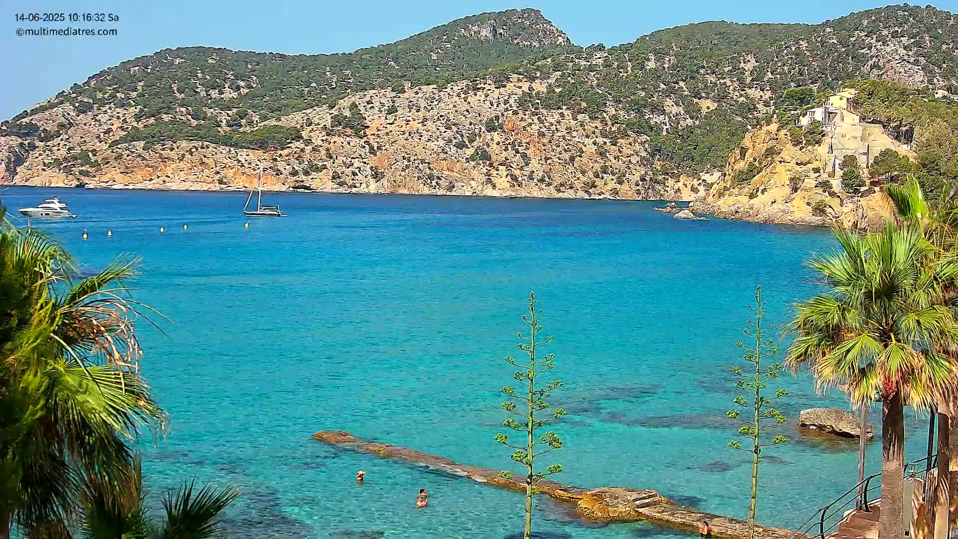 A picturesque cove with clear turquoise water, two boats, and palm trees framing the scene under a sunny sky.