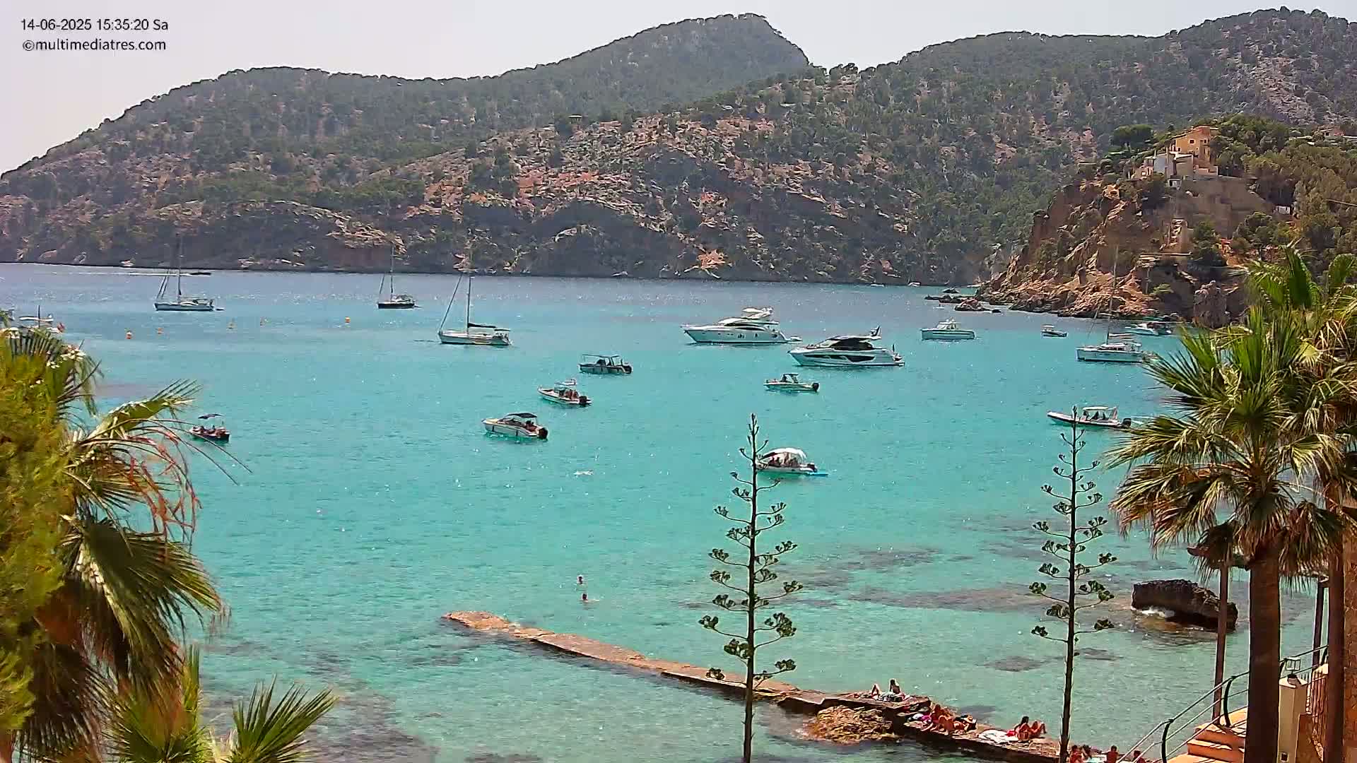 Several boats are anchored in a calm, turquoise bay surrounded by rocky hills and palm trees on a sunny day.