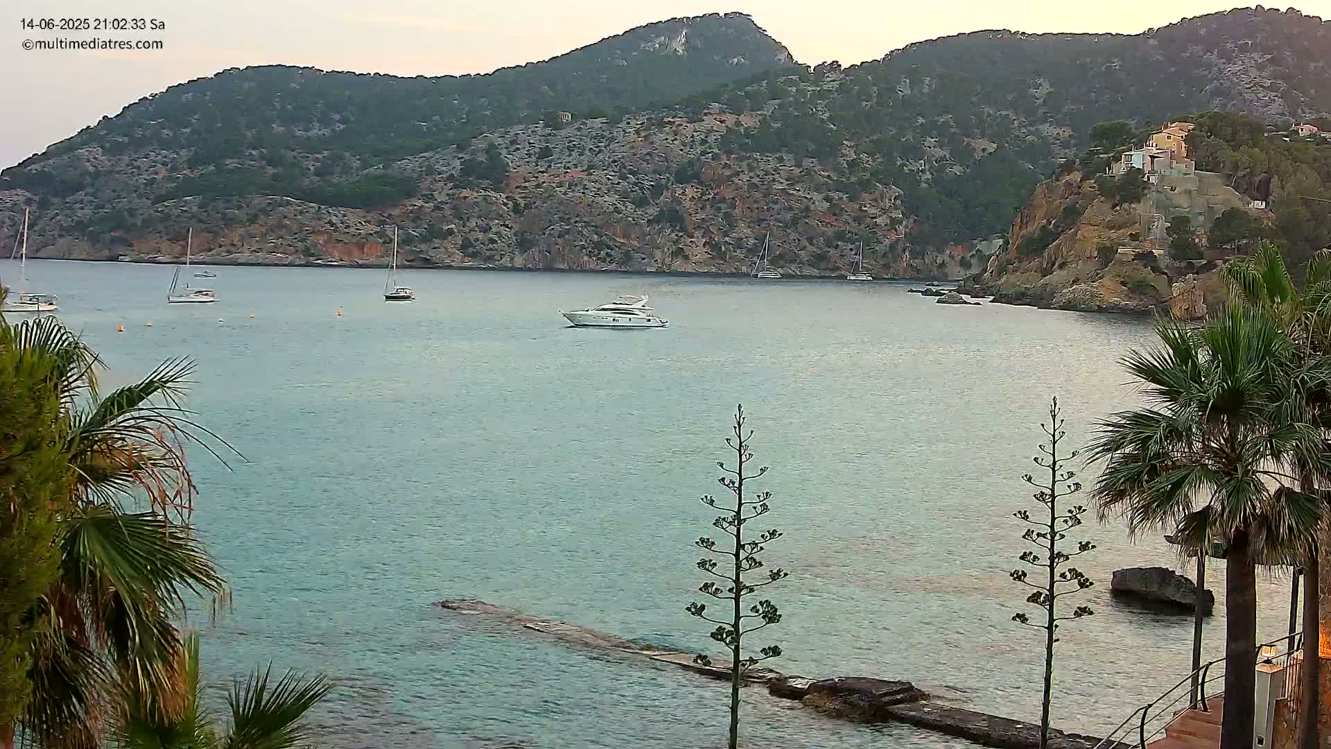 Several boats are anchored in a calm, turquoise bay surrounded by rocky hills and palm trees on a sunny day.