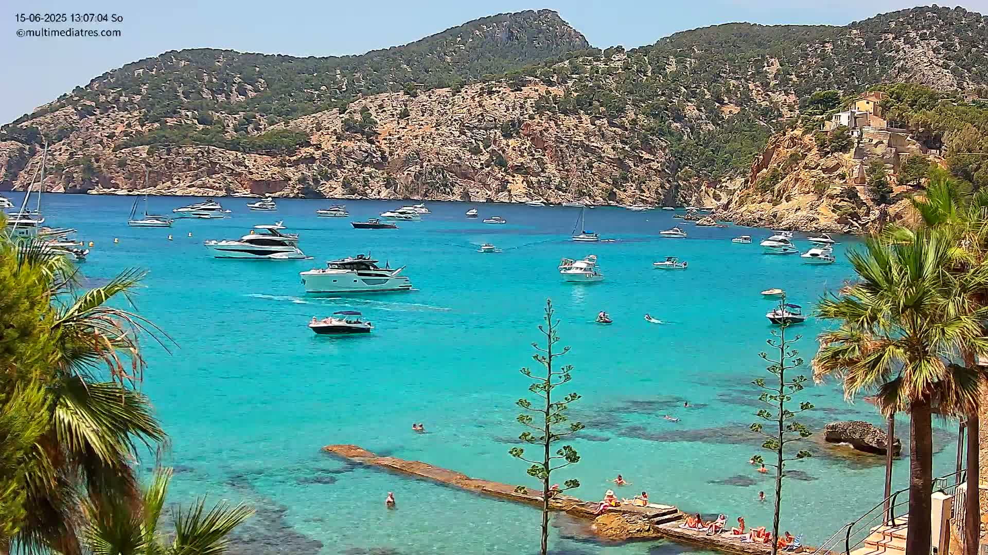 A bright turquoise cove, surrounded by rocky hillsides and palm trees, is filled with numerous boats and people swimming on a sunny day.