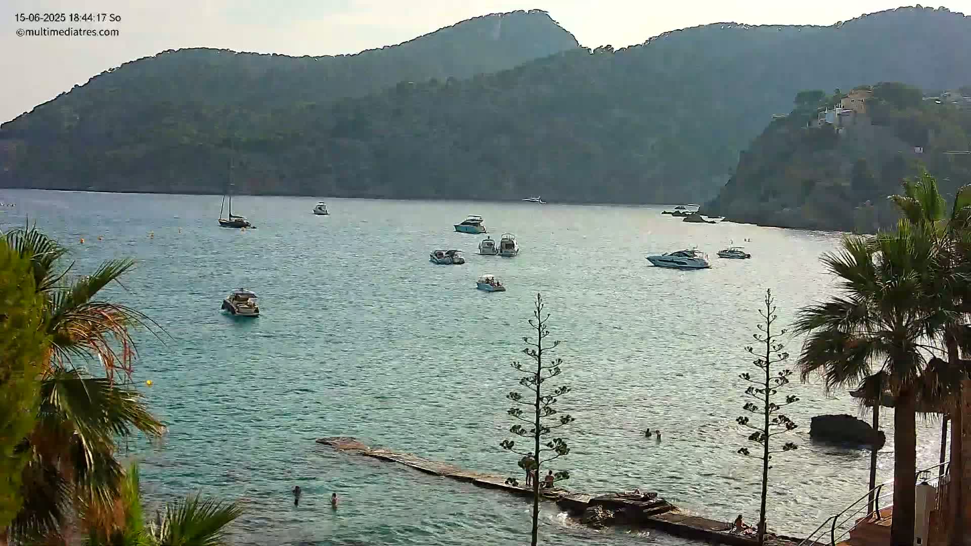 A calm, clear body of water is filled with several boats near a rocky shoreline, backed by green hills under a hazy sky.