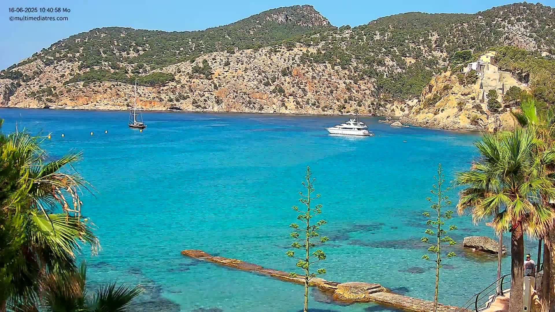 A bright, sunny day shows a tranquil cove with clear, turquoise water, two boats, and palm trees lining a rocky shore, backed by a verdant hillside.