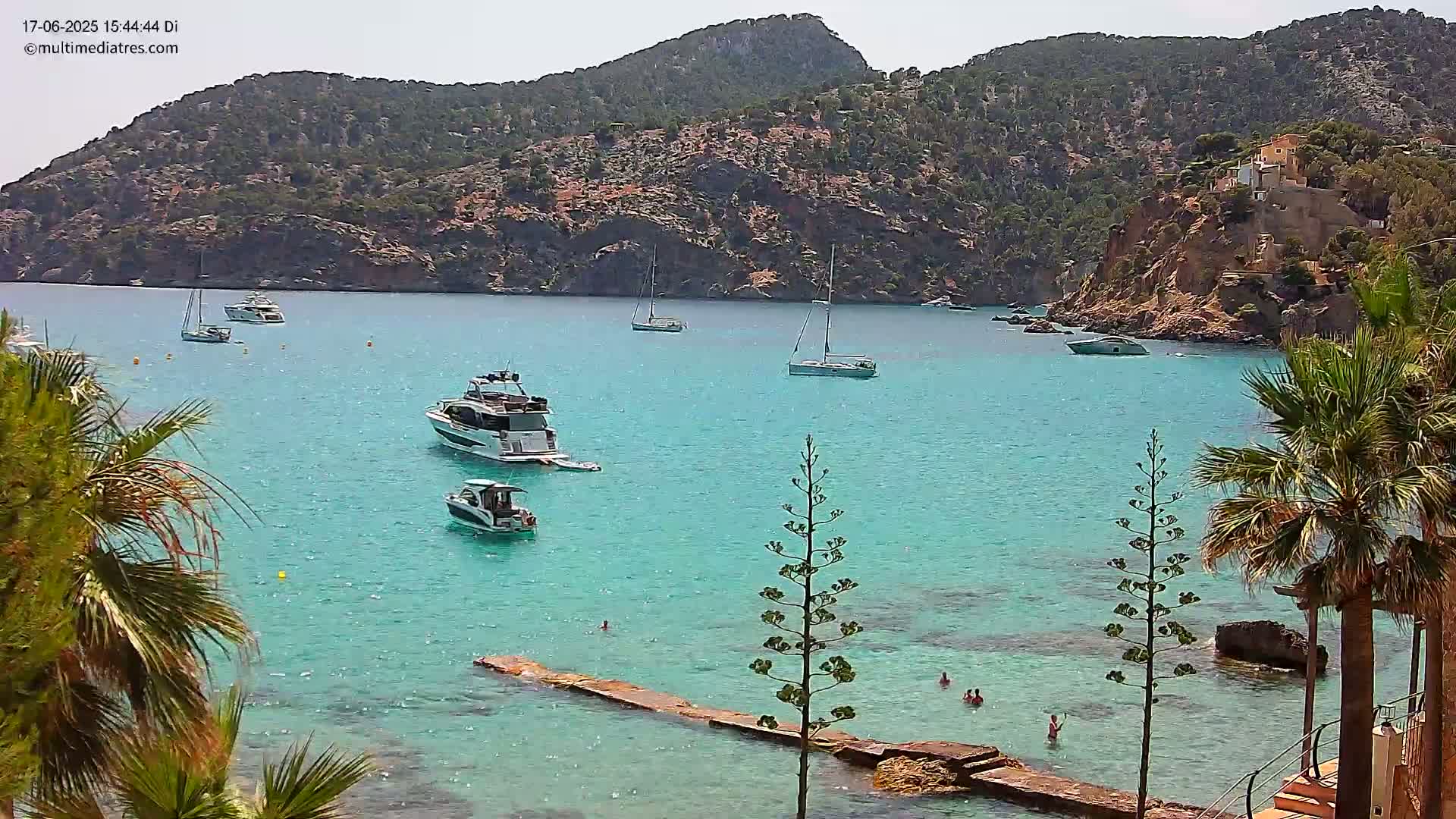 Several boats are anchored in a calm, turquoise bay surrounded by rocky hillsides and palm trees on a sunny day.