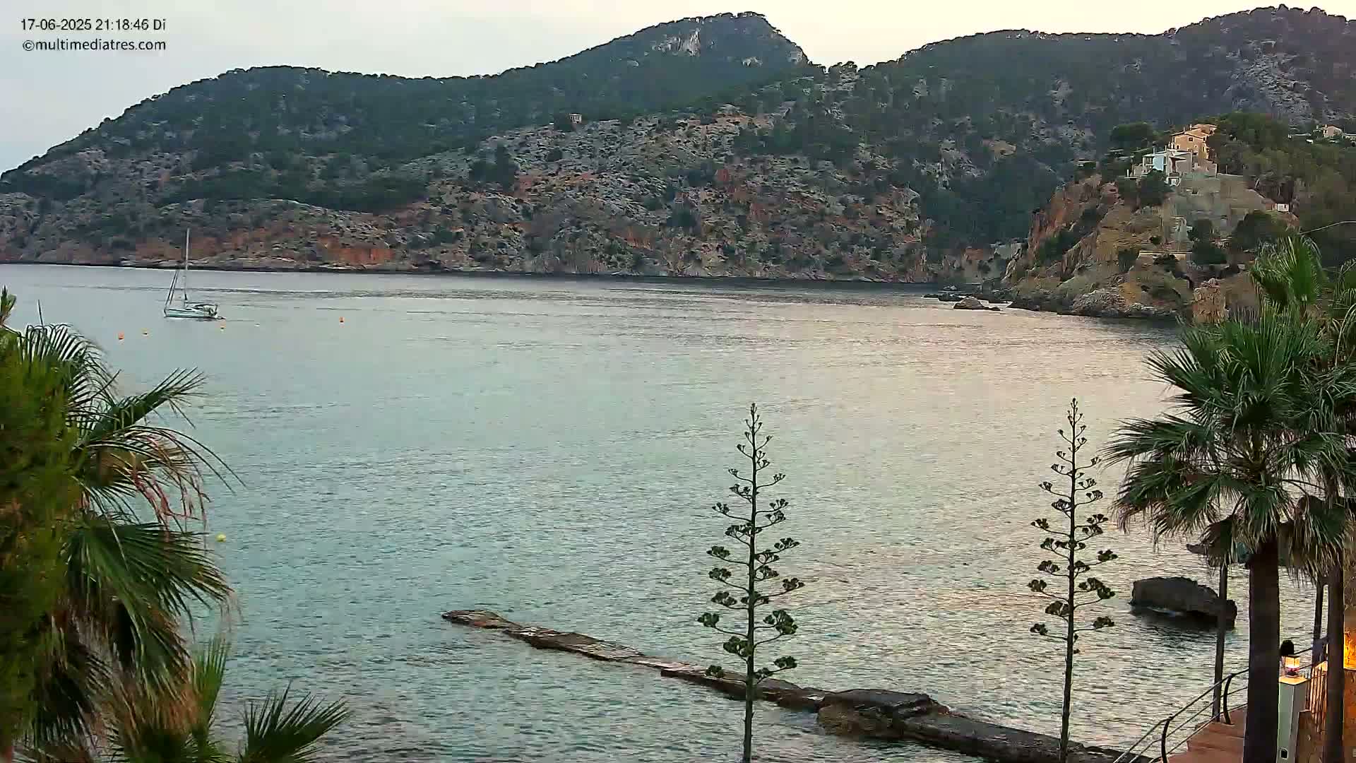 A sailboat is visible on calm, light-green water near a rocky shoreline with vegetation, including palm trees, under a mostly clear sky.