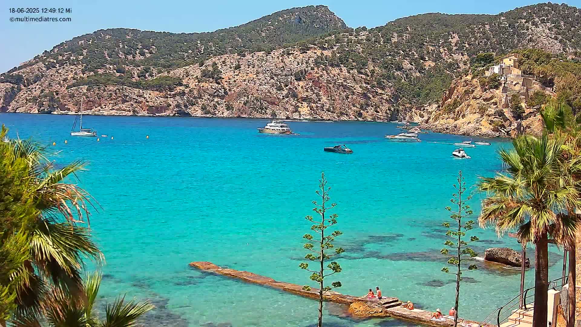A picturesque, sunny cove with clear turquoise water, several boats, and people relaxing on a stone jetty near palm trees.