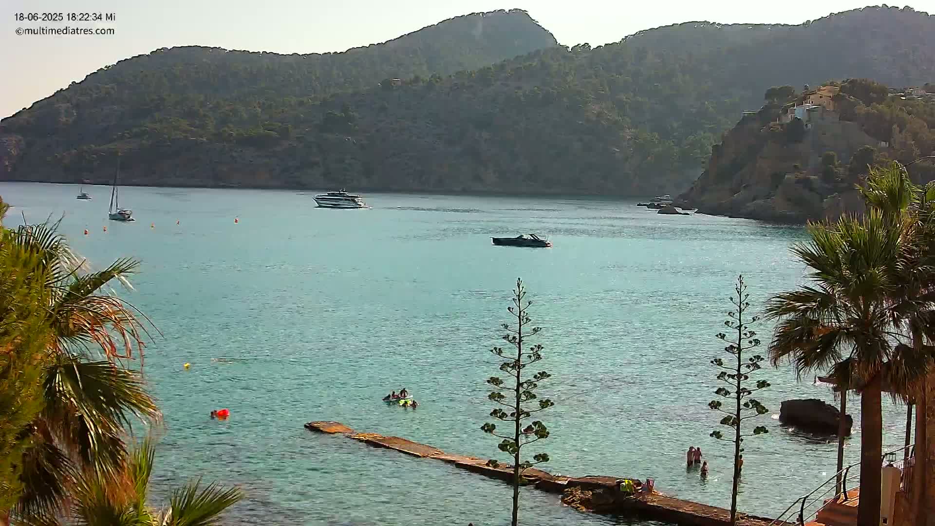 A calm, clear turquoise bay is surrounded by green hills, with several boats and people swimming in the water near a small wooden dock, under a bright, sunny sky.