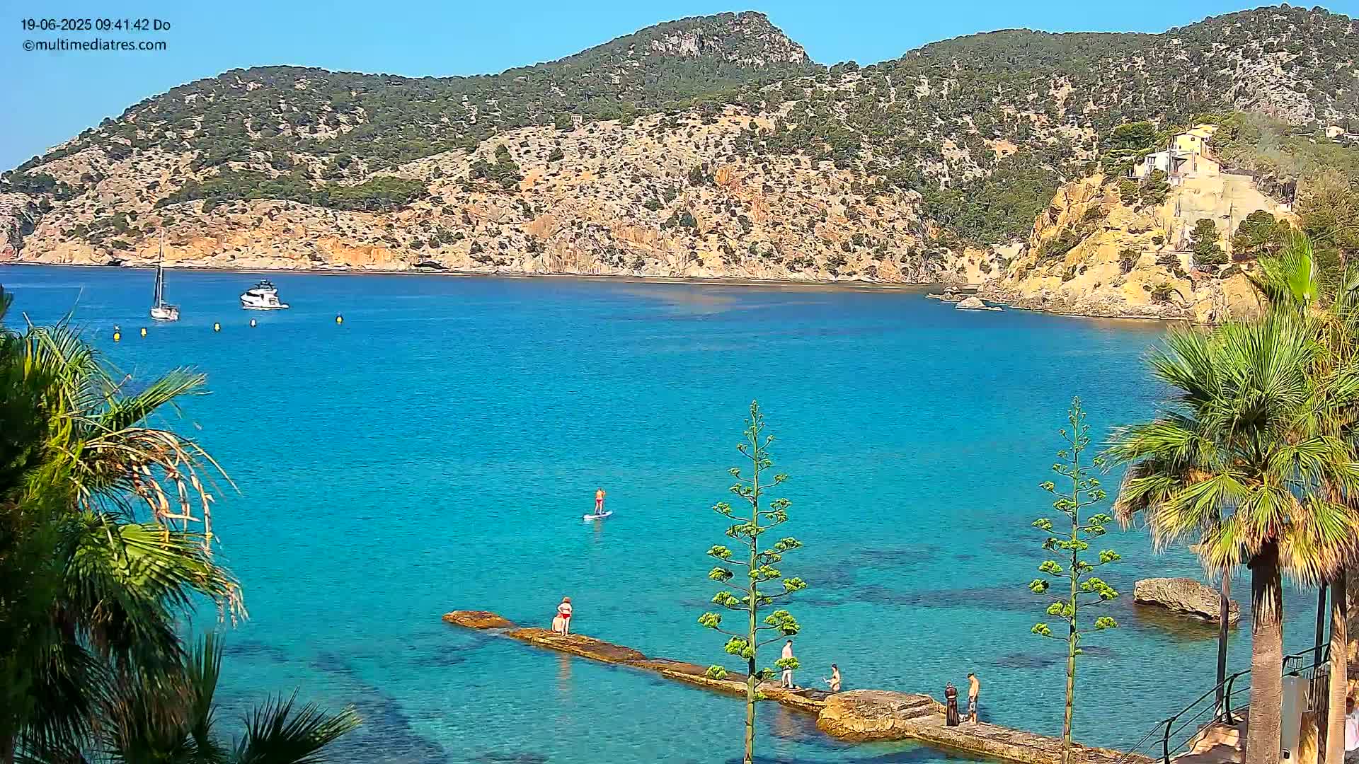 A calm, bright blue bay with two boats, several people on rocks in the water, and palm trees in the foreground under a clear sky.