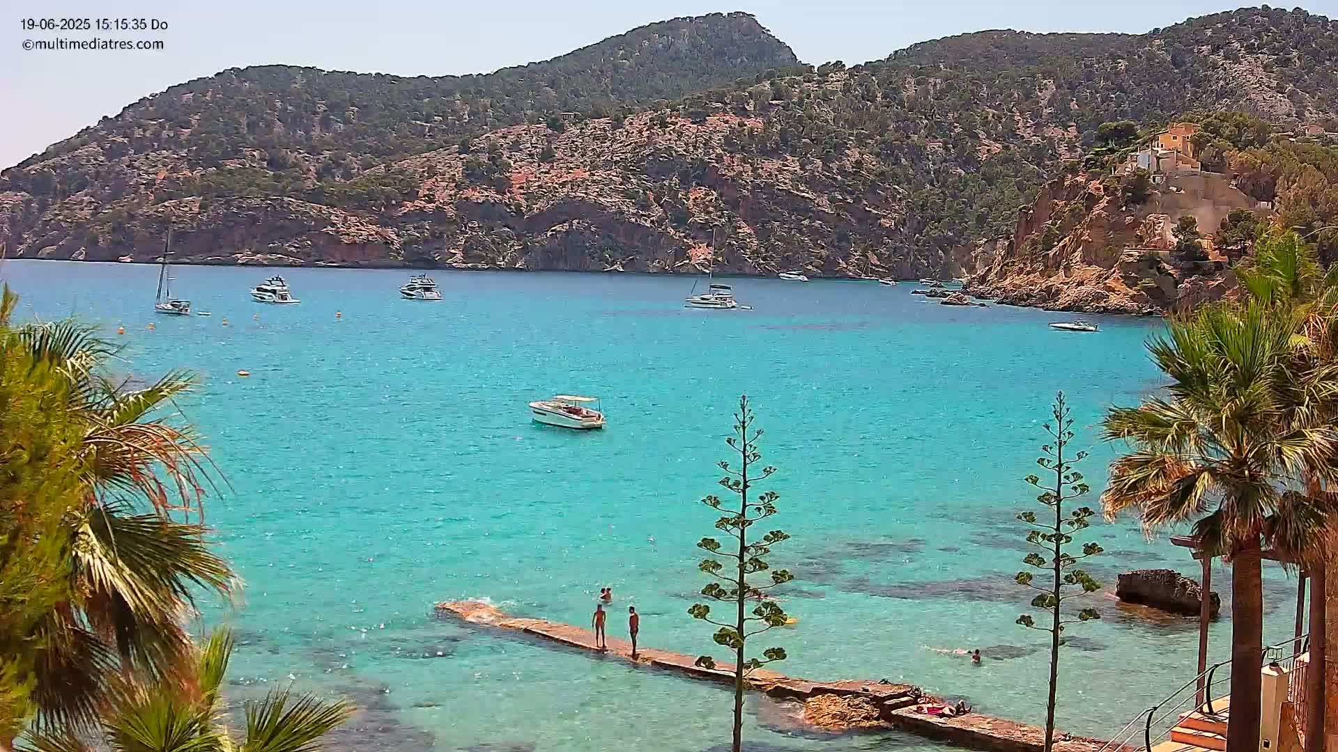 A clear, sunny day reveals a turquoise bay with several boats anchored near a rocky, hilly coastline, where people relax on a small jetty in shallow water.
