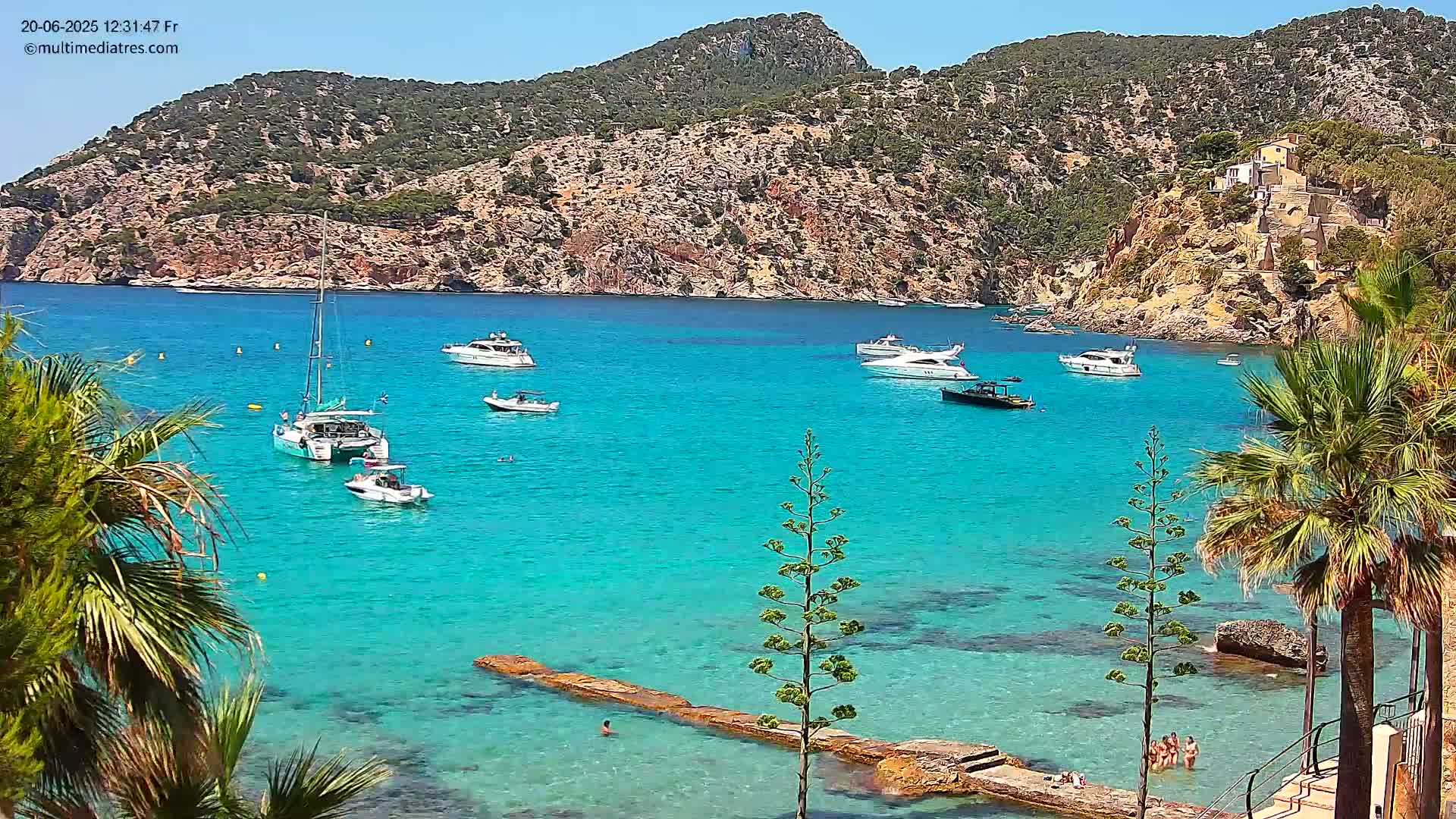 Several boats are anchored in a calm, turquoise bay, surrounded by a rocky hillside with sparse vegetation under a clear, sunny sky.