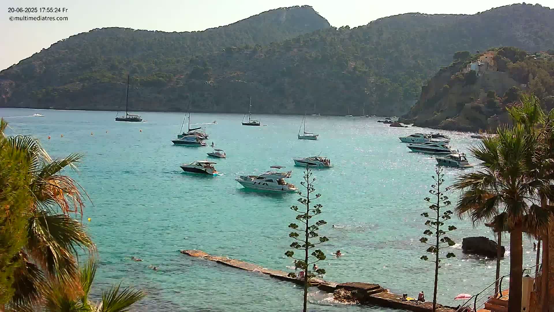 Numerous boats are anchored in a calm, turquoise bay, surrounded by palm trees and rocky hills under a bright, sunny sky.
