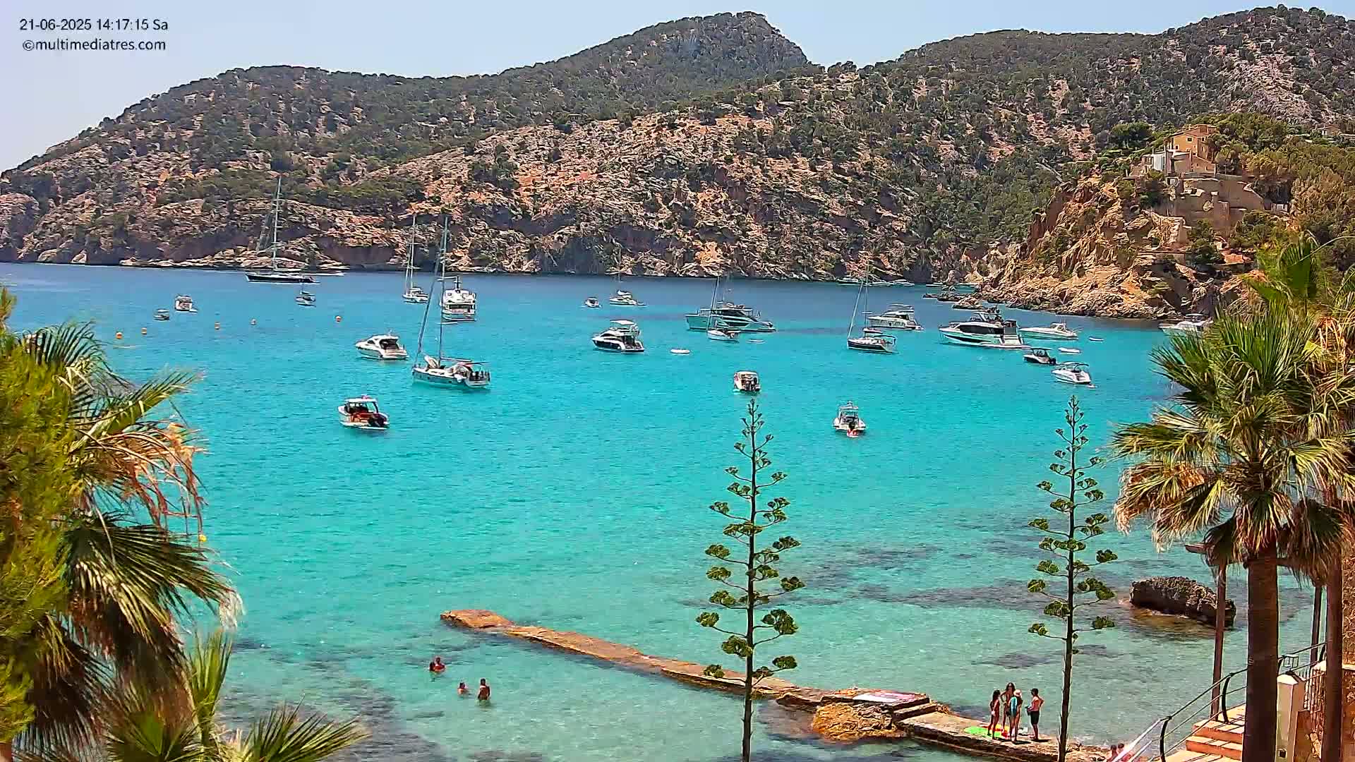 A bright, sunny day shows a calm, turquoise bay with numerous boats anchored, surrounded by rocky hillsides with sparse vegetation and palm trees.