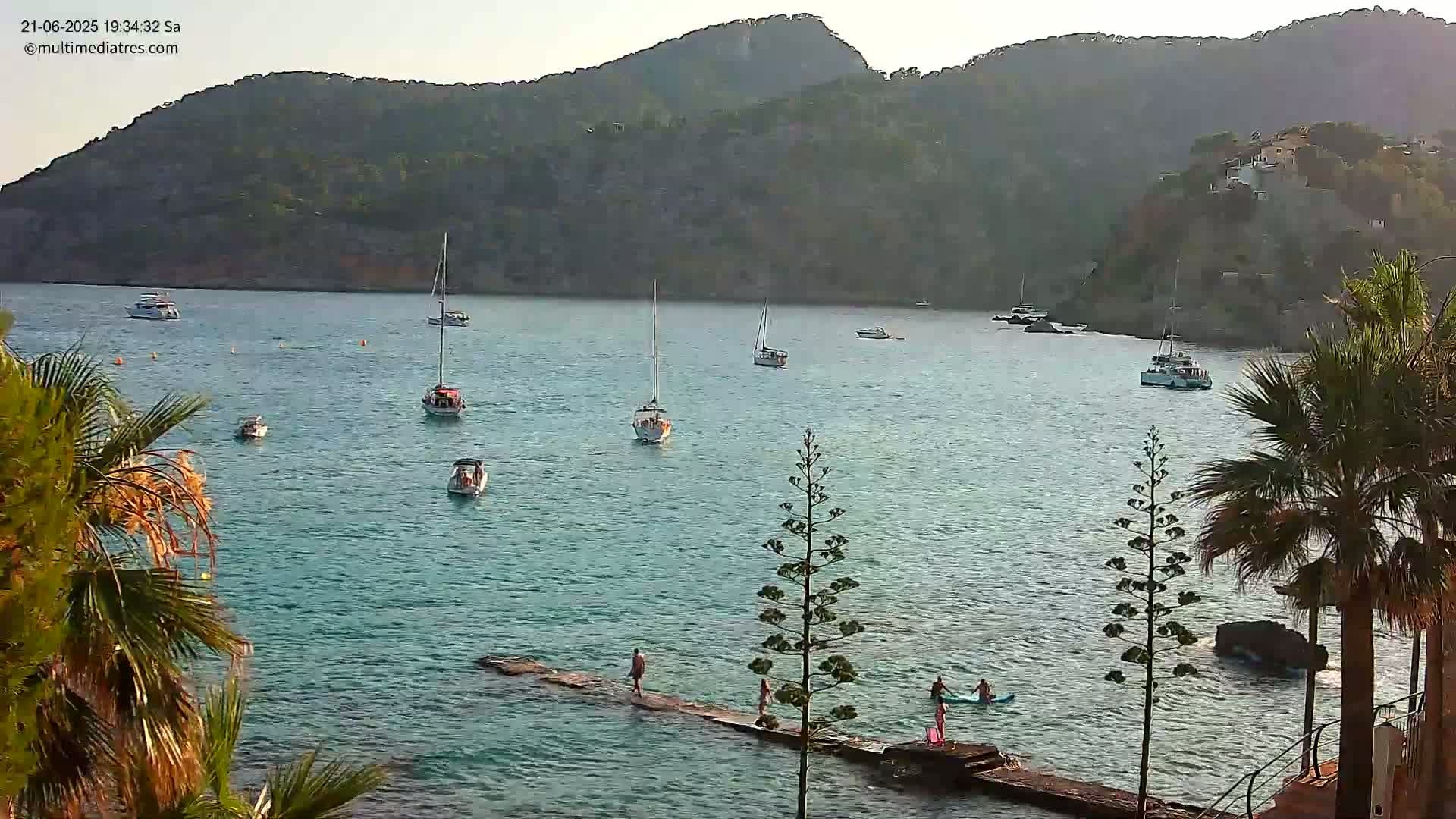 A calm, clear day shows several boats in a bay surrounded by hills, with people on a jetty and in the water near palm trees.