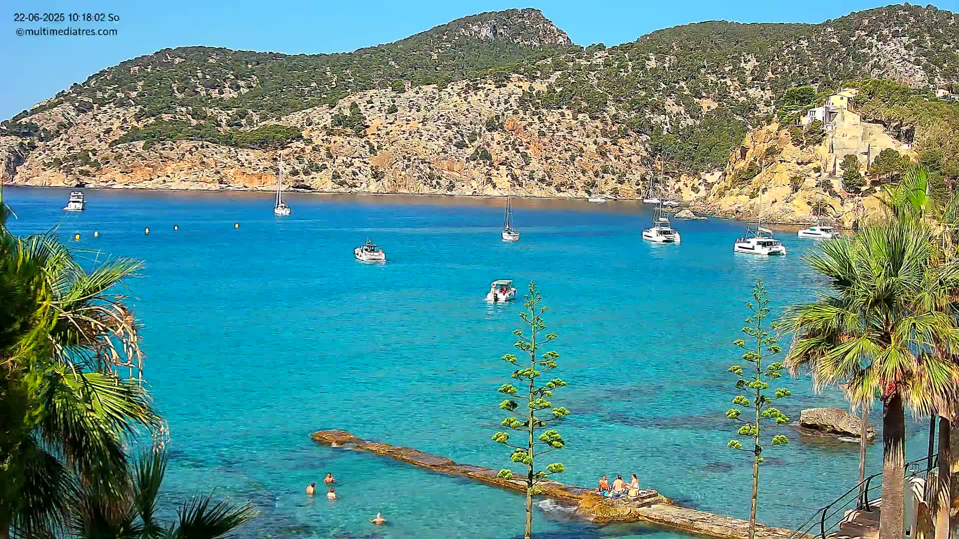 A sunny, clear day shows a calm, bright turquoise bay with several boats and people swimming and relaxing on rocks near palm trees and a rocky hillside.