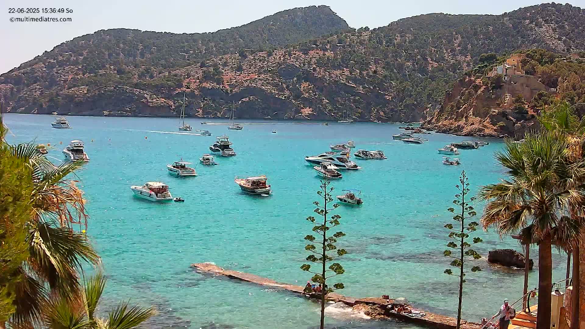 Numerous boats are anchored in a calm, turquoise bay surrounded by a rocky, green hillside under a clear, sunny sky.