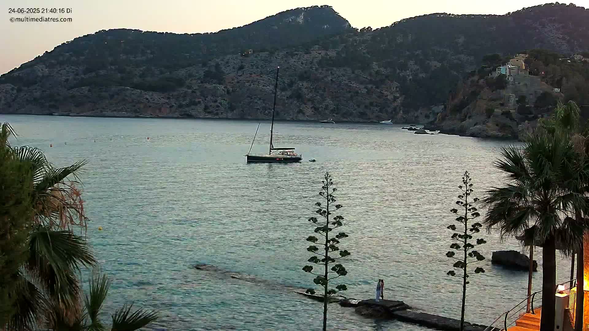 A sailboat is anchored in a calm, twilight bay surrounded by dark hills and palm trees, with a couple standing on a rock jetty in the foreground.