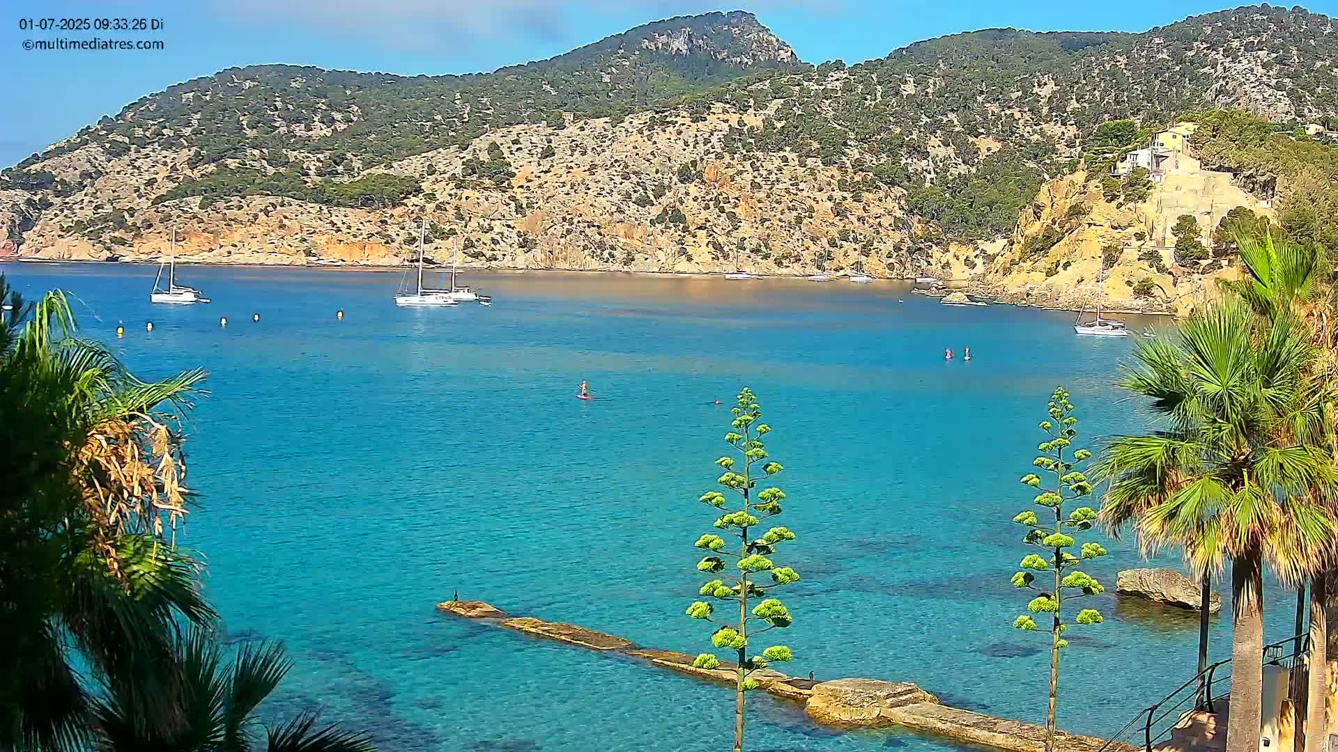 A tranquil turquoise bay is nestled between verdant hills under a clear sky, with several sailboats at anchor and a person paddleboarding in the calm water.