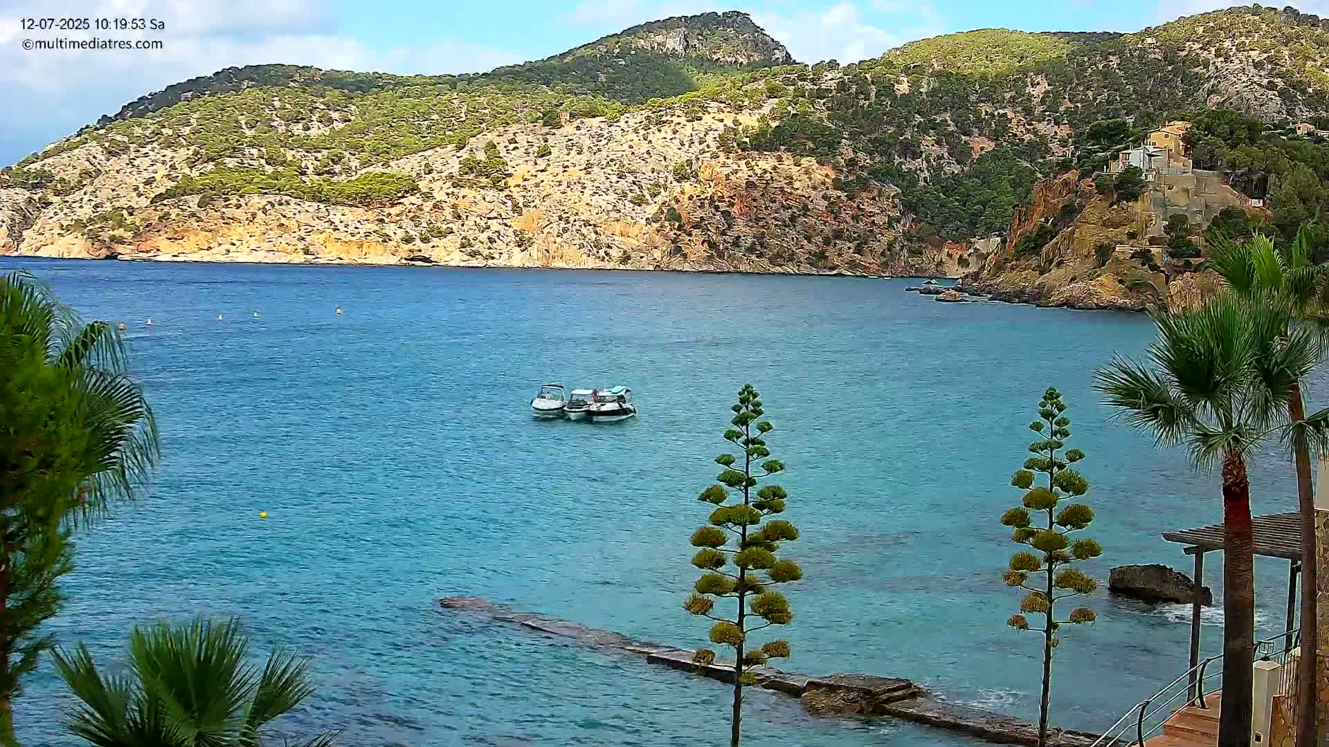 Two small boats are anchored in a calm, clear turquoise bay nestled against a rocky, green-covered hillside under a mostly sunny sky.