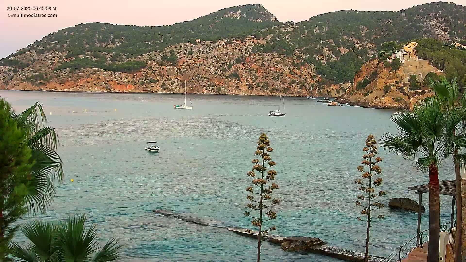 A calm, blue ocean with gentle waves laps against a rocky shore, where several palm trees stand near a small structure, with boats visible in the distance and a hilly, green island in the background under a partly cloudy sky.