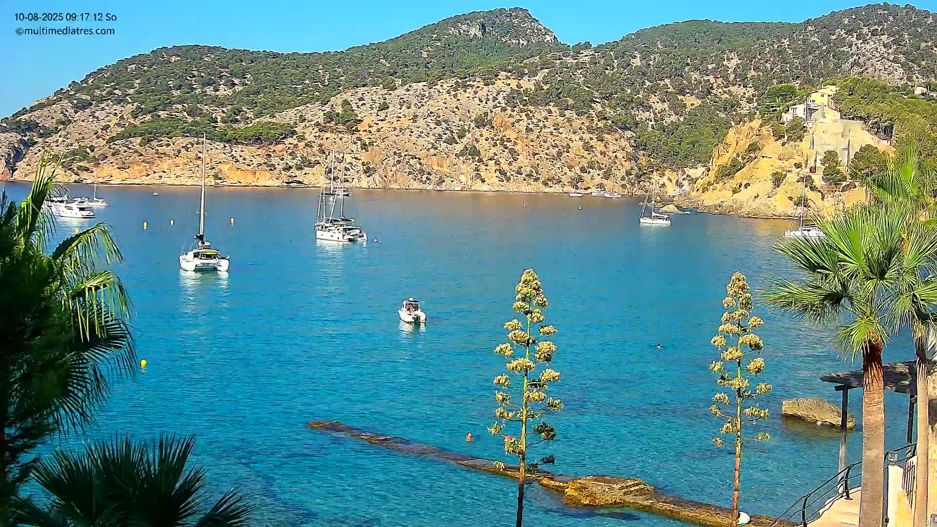 A calm, sunny day shows a clear blue bay with several sailboats and motorboats anchored near a rocky, green hillside dotted with buildings.