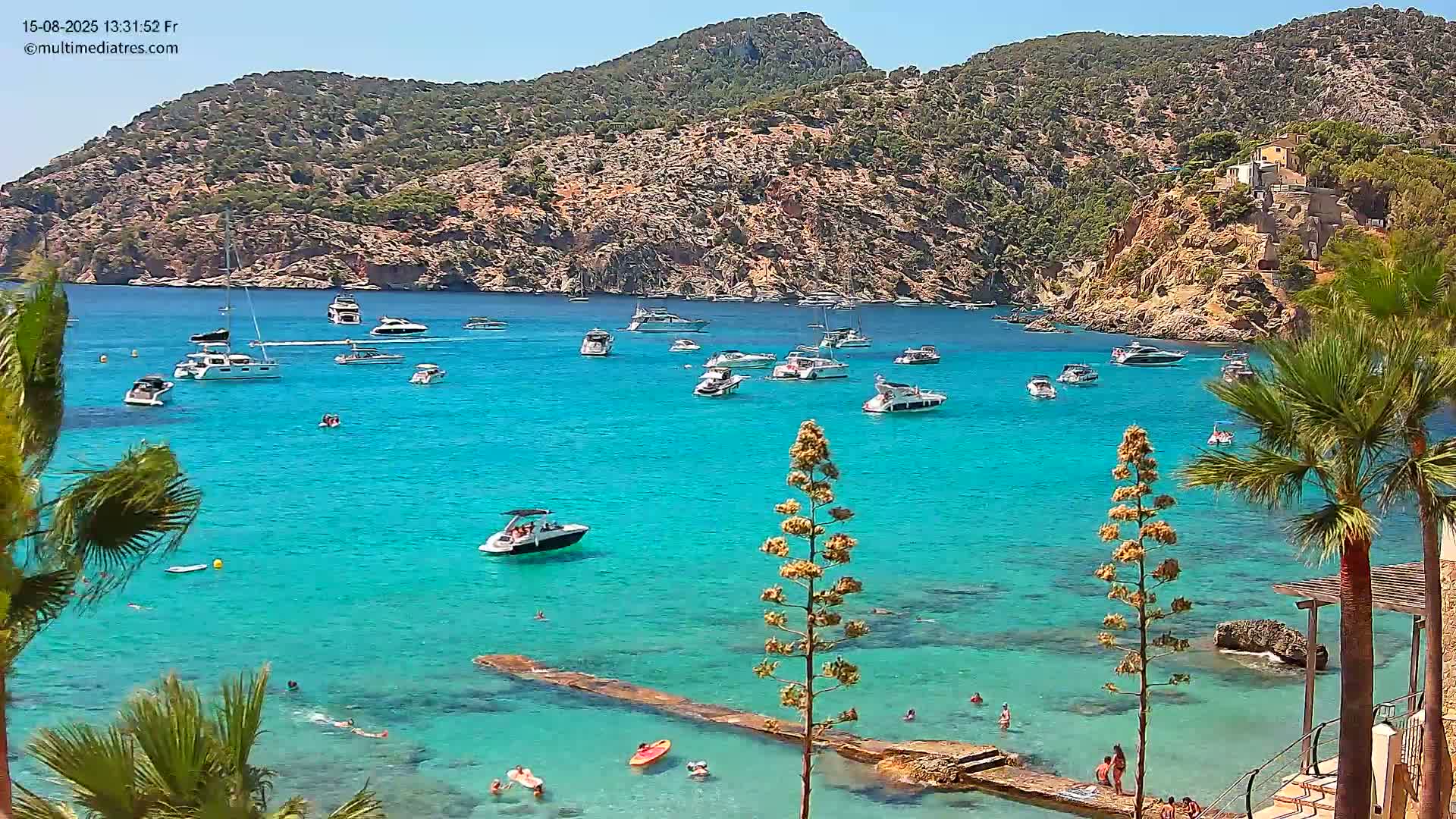 A calm, sunny day shows a clear blue bay with several sailboats and motorboats anchored near a rocky, green hillside dotted with buildings.