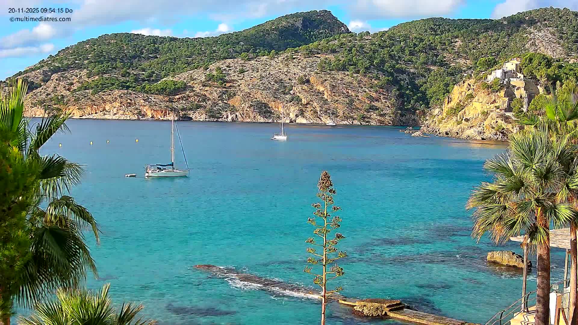 A clear, sunny day reveals a vibrant turquoise bay dotted with several sailboats, framed by rocky, tree-covered hills and coastal vegetation, with calm waters.