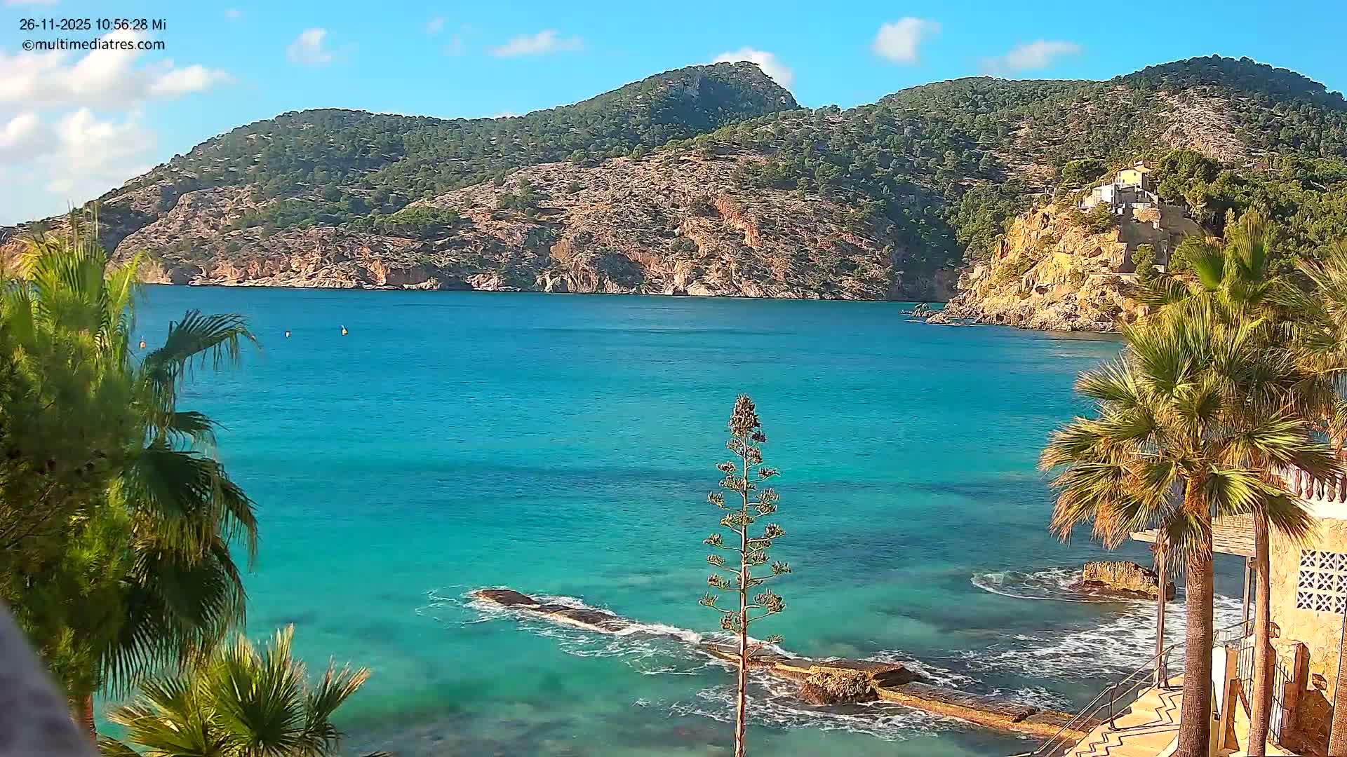 A vibrant turquoise bay is nestled between steep, green-covered hills under a clear blue sky with scattered clouds on a sunny day, with palm trees and a building complex on a cliff framing the foreground.