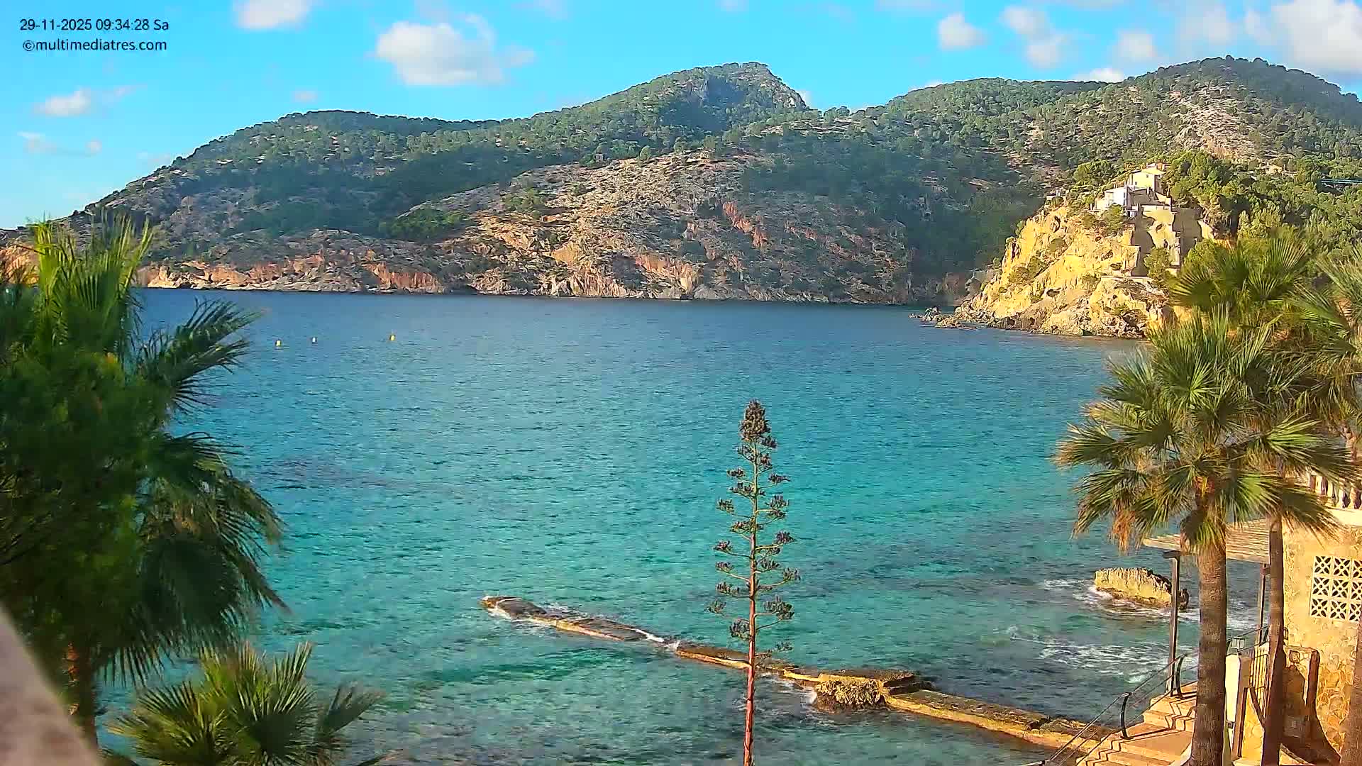 On a bright, sunny day with a partly cloudy blue sky, a tranquil turquoise bay is framed by steep, forested hills and a rocky coastline, with palm trees and a submerged rock structure in the foreground.