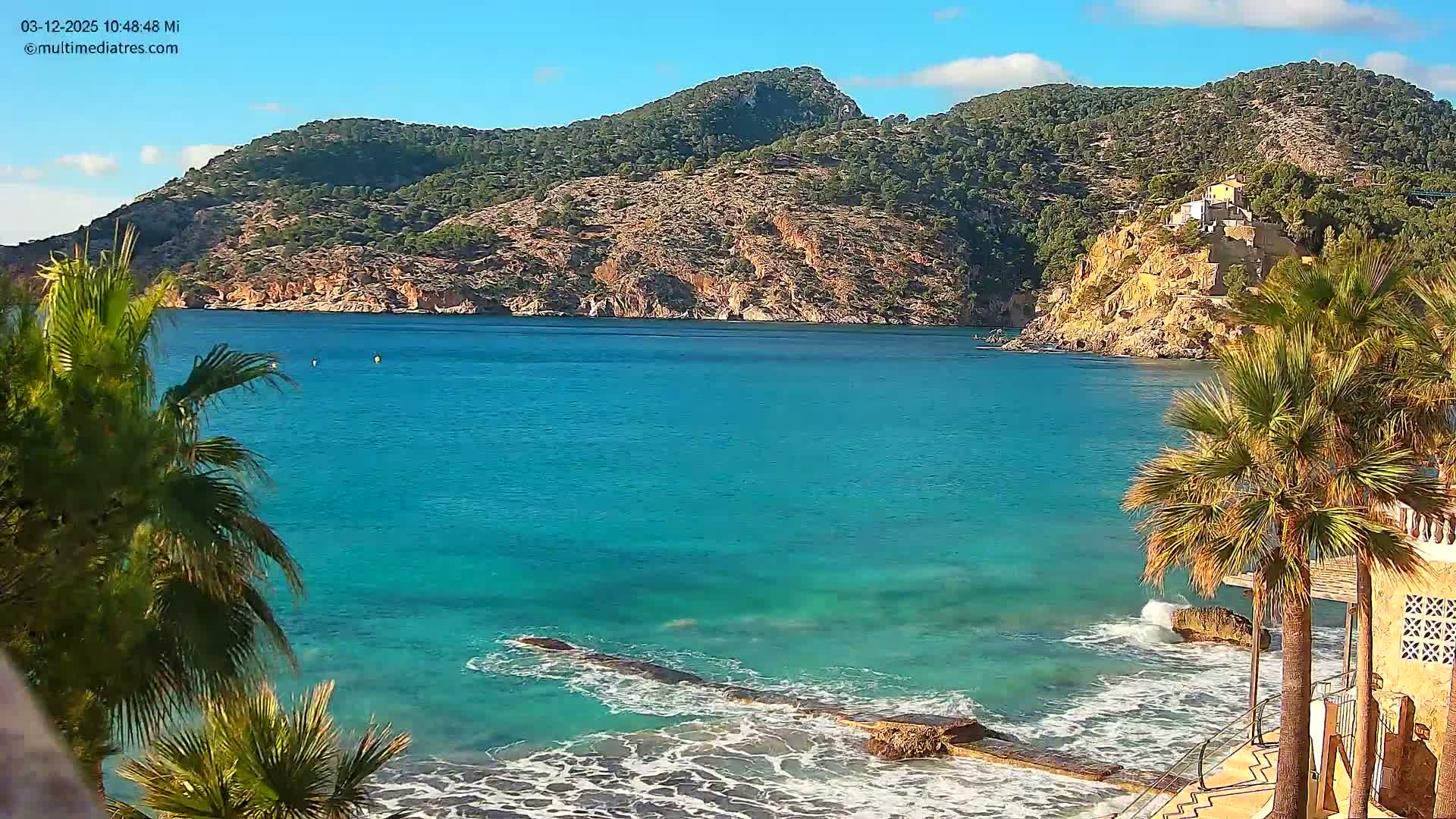 The image shows a vibrant turquoise bay with gentle waves lapping a rocky shore in the foreground, framed by palm trees, with lush green hills and a small building perched on a distant cliff under a sunny blue sky with scattered white clouds.