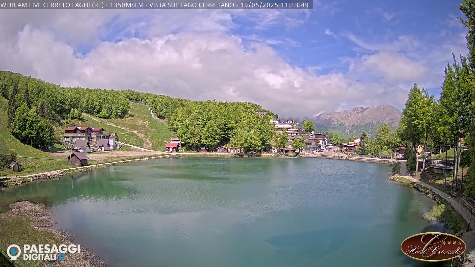 A tranquil lake surrounded by lush green hills and mountains under a partly cloudy sky is shown.