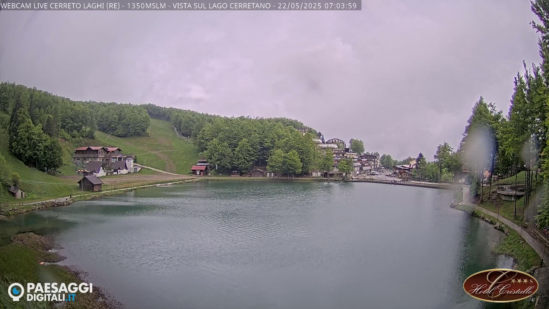 A calm lake sits before a small village nestled in a green, hilly landscape under an overcast sky.