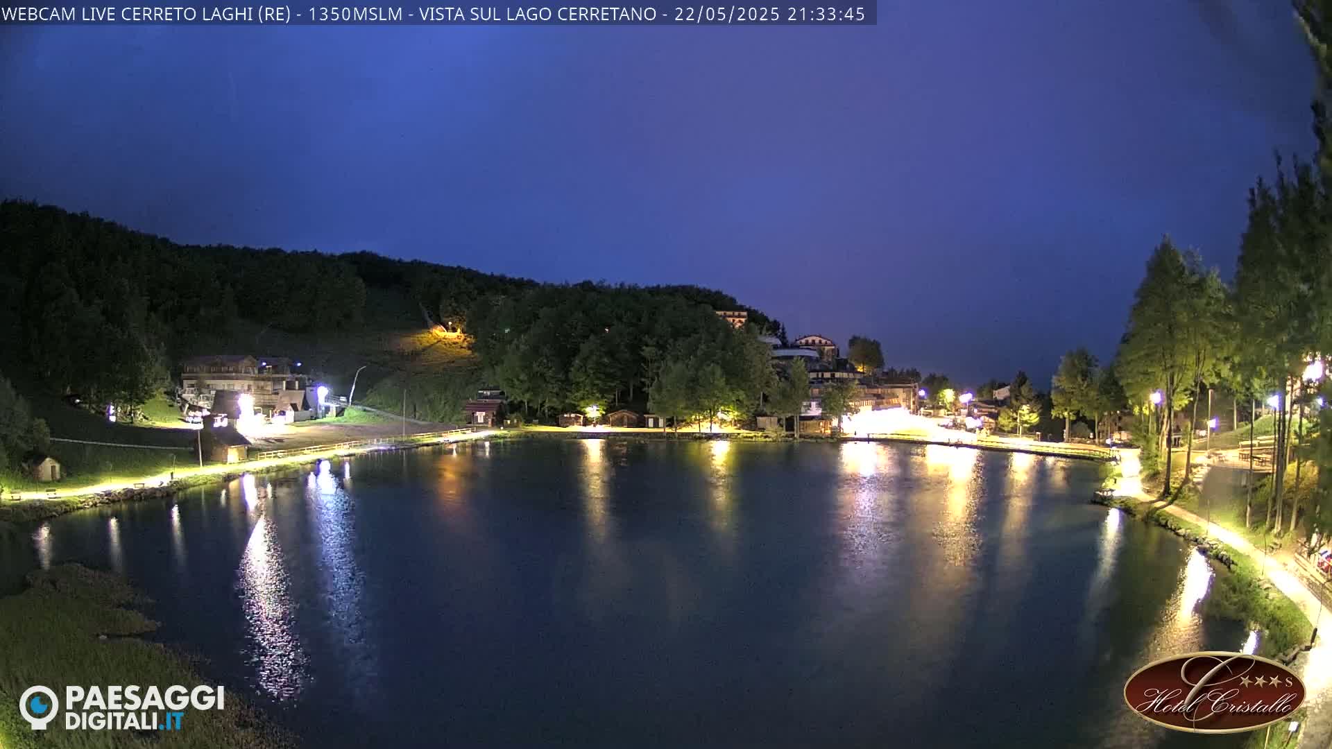 A nighttime view of a calm lake reflecting illuminated buildings and trees under a twilight sky.