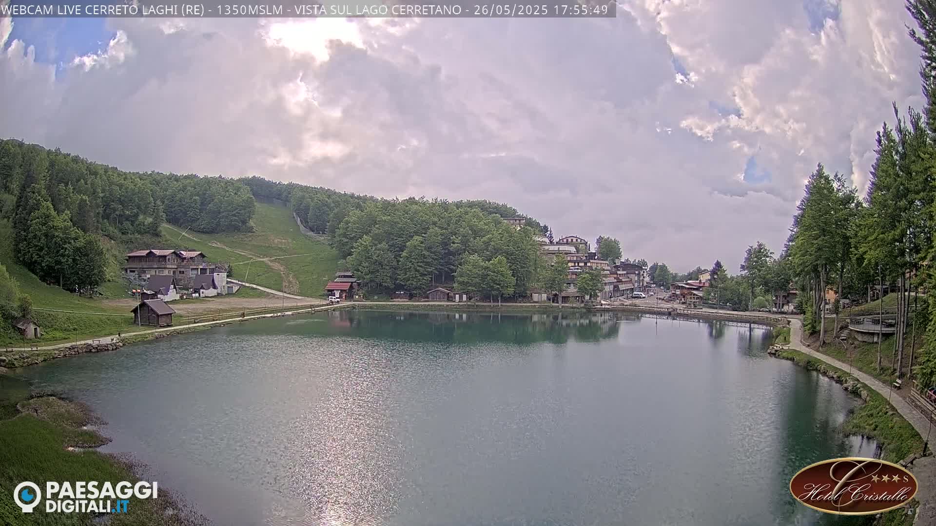 A calm lake reflects a village nestled amongst green hills under a partly cloudy sky.
