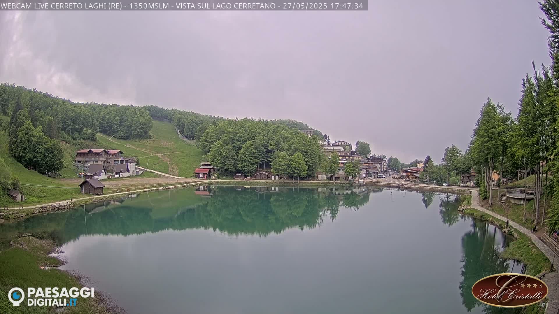 A calm lake reflects buildings and trees under an overcast sky.