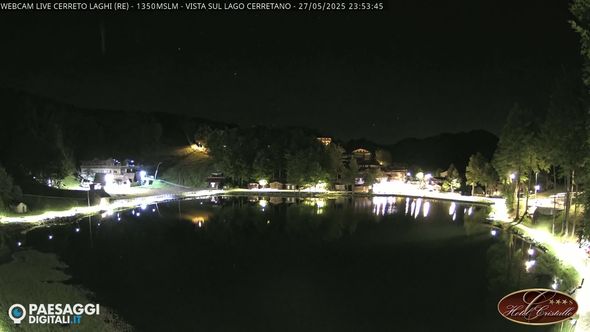 A nighttime view of a calm lake reflecting the lights of buildings and trees on its shores under a clear, dark sky.