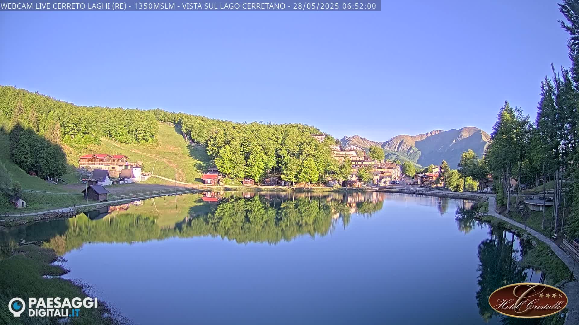 A calm lake reflects a village nestled among green hills and mountains under a clear blue sky.
