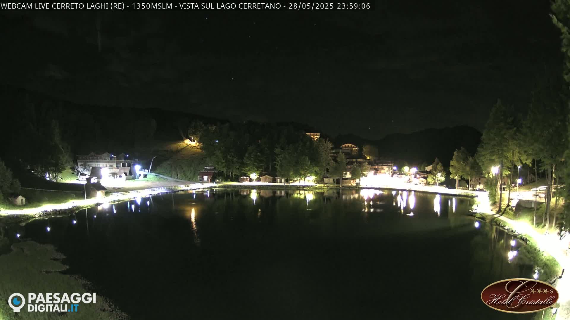 A calm lake at night reflects the lights of a village nestled among dark hills under a clear sky.