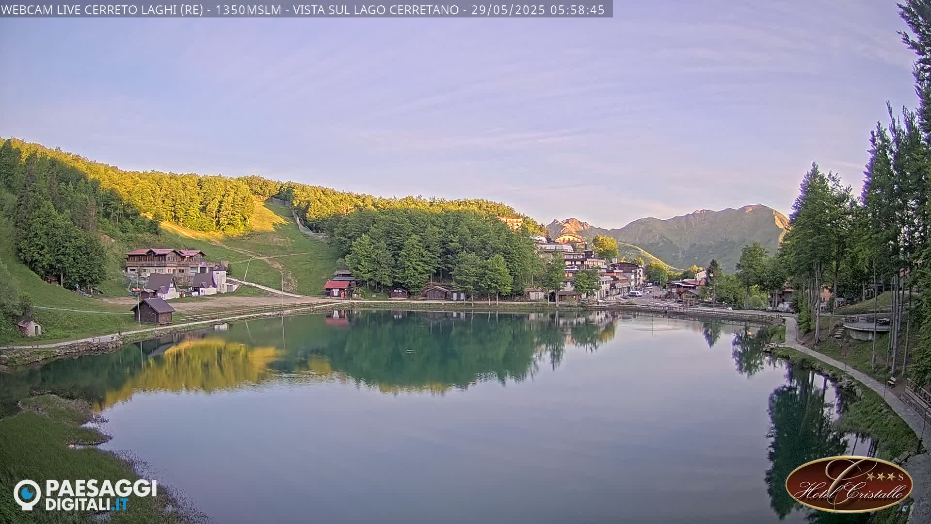A calm lake reflects a small village nestled among green hills and mountains under a clear, light-blue sky.