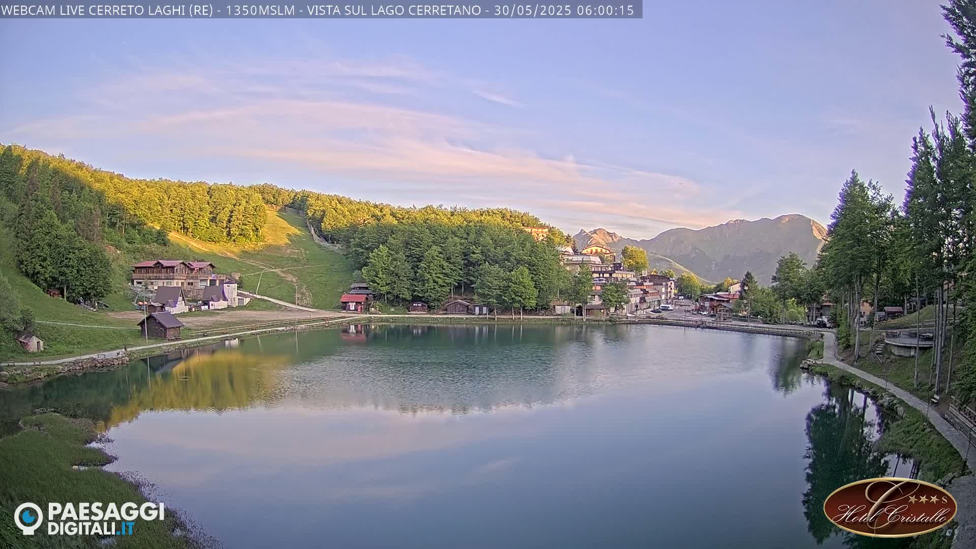 A calm lake reflects a small village nestled among green hills and mountains under a clear, sunrise sky.