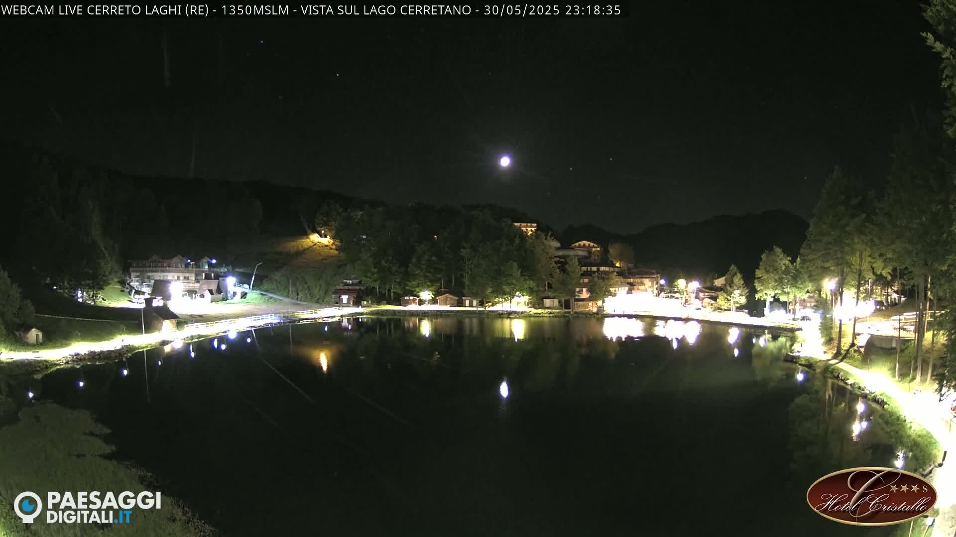 A moonlit night scene shows a calm lake reflecting illuminated buildings and trees along its shore.