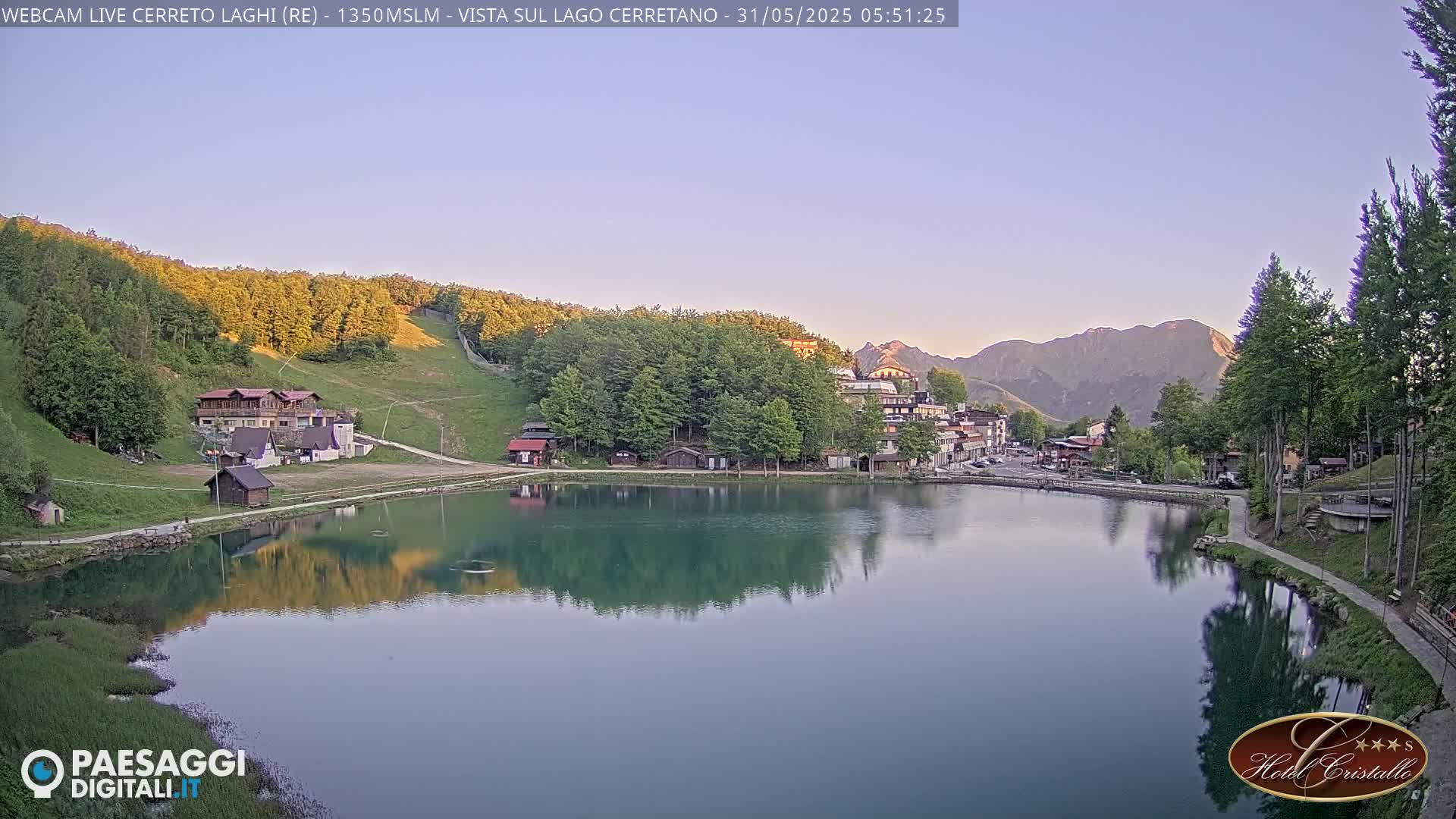 A calm lake reflects a village nestled in a mountain valley at dawn under a clear sky.